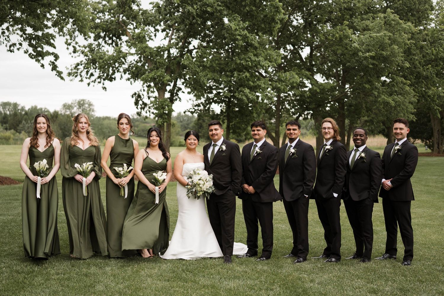 Wedding party in sage green bridesmaid dresses and dark suits posing on green lawn.
