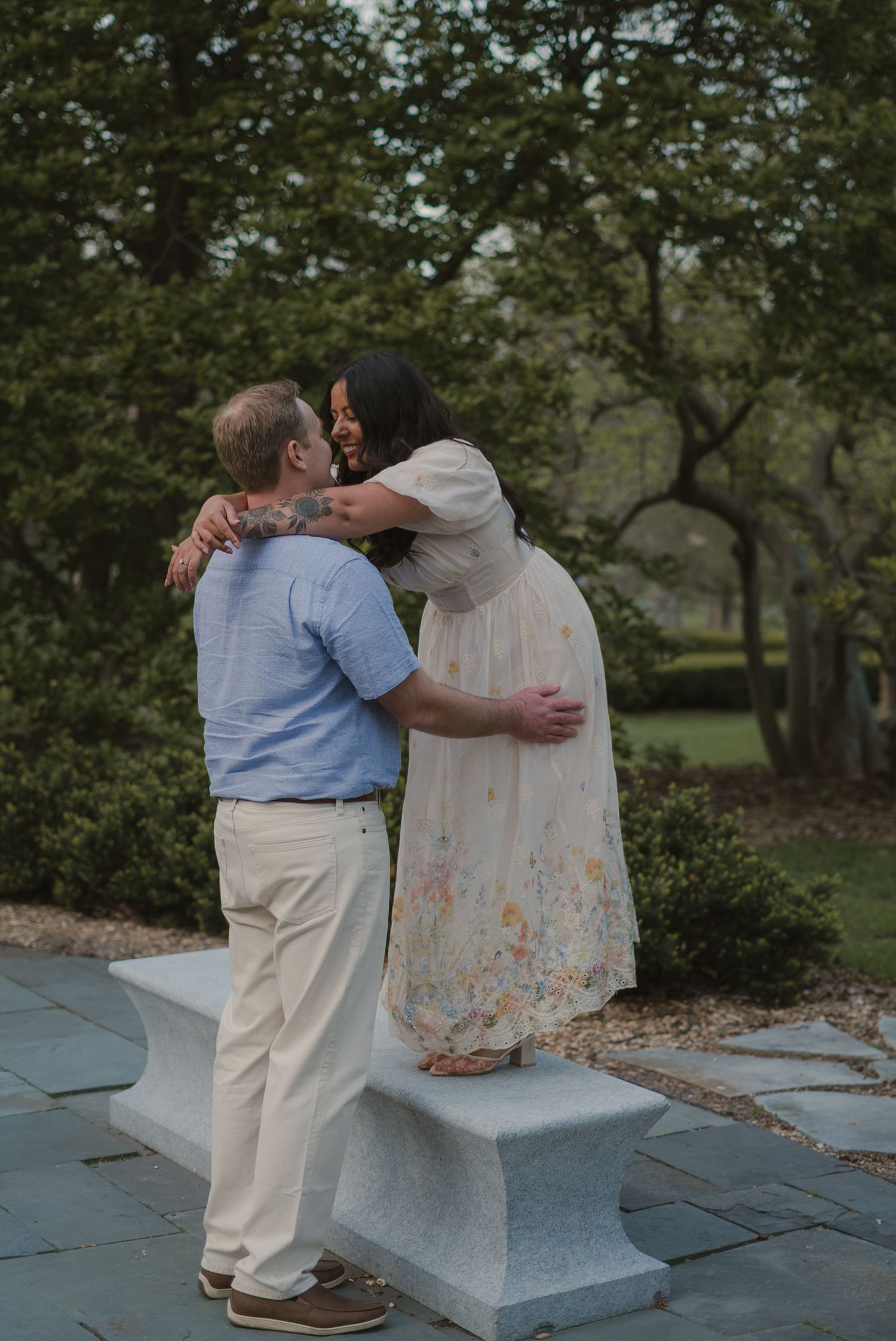 Couple embracing and kissing in a garden, woman in floral dress standing on stone bench, man in blue shirt.