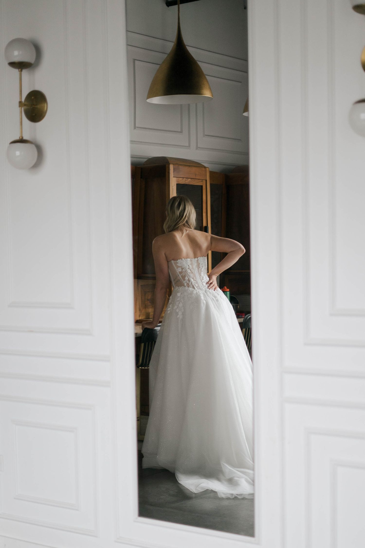Bride in a strapless white ball gown wedding dress viewed from behind through a doorway in an elegant room.