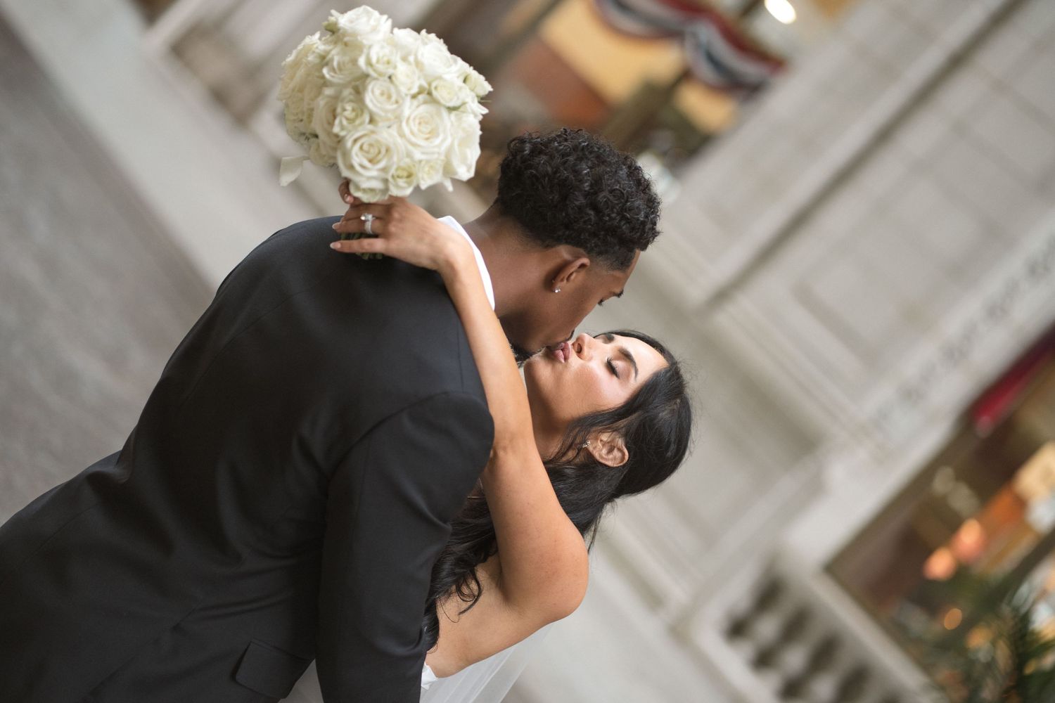 A bride and groom share a romantic kiss, with the bride holding a white rose bouquet at their wedding.