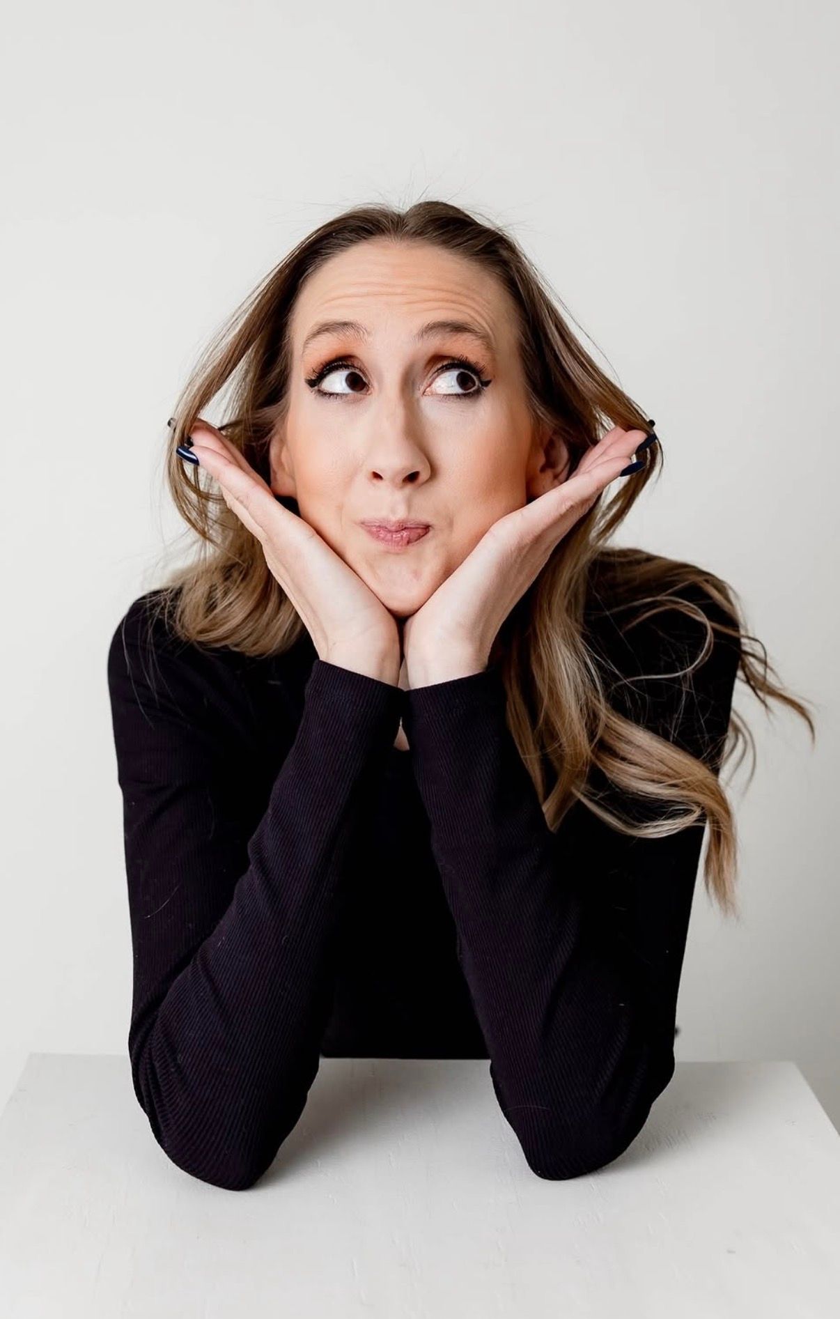 Woman in black top rests chin on hands, making a playful expression while looking up against white background.