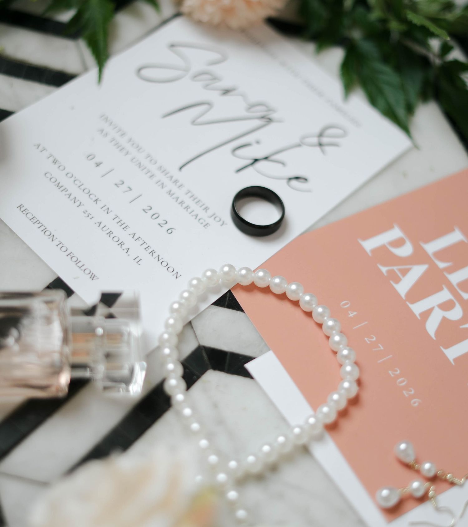 Elegant wedding flat lay with pearl bracelet, black ring, floral invitations, and perfume bottle on striped surface.