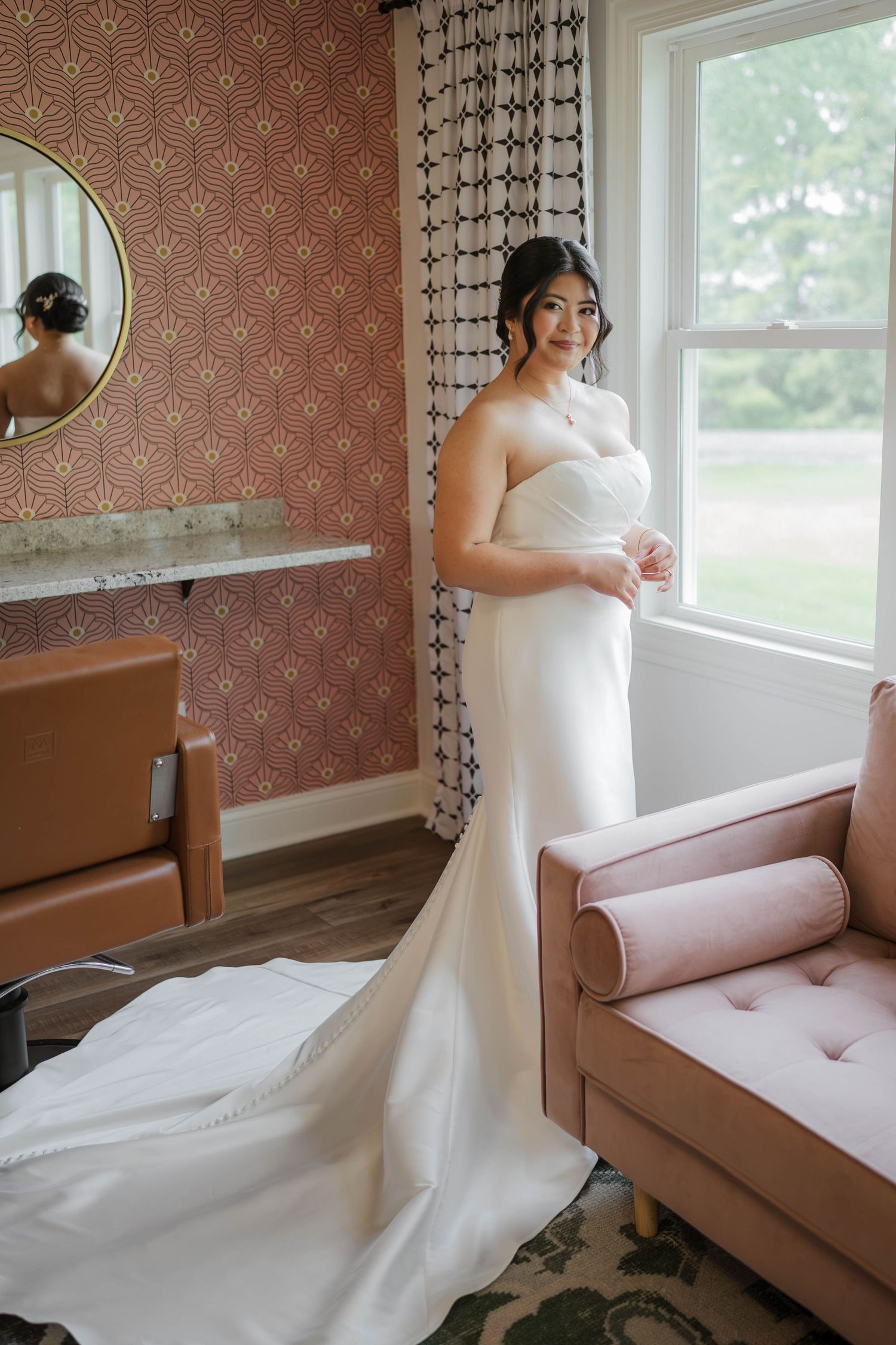 Bride in a strapless white mermaid wedding gown standing by a window in an elegant pink-walled bridal suite.