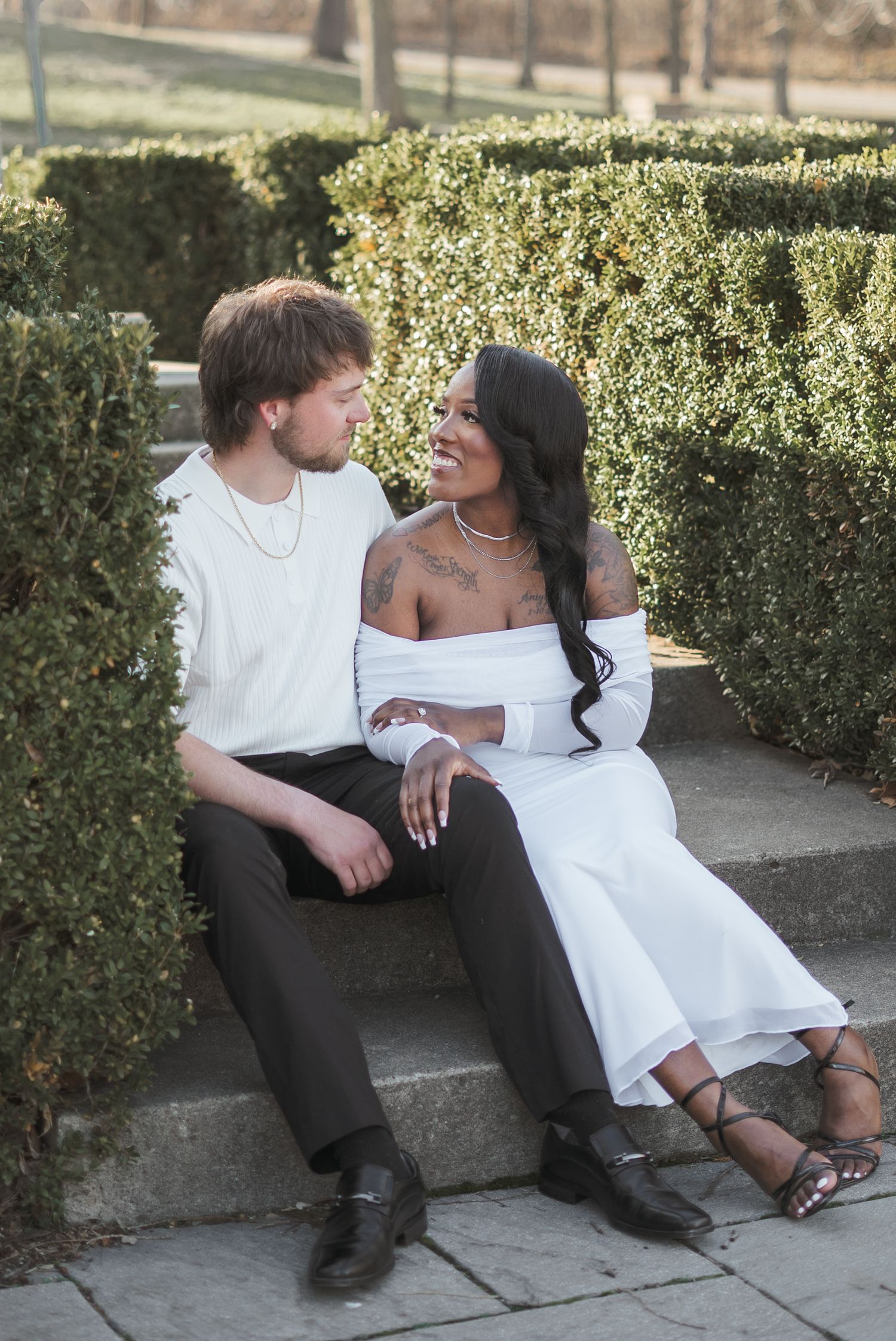 Interracial couple sitting on stone steps in a garden, smiling at each other, wearing white outfits.
