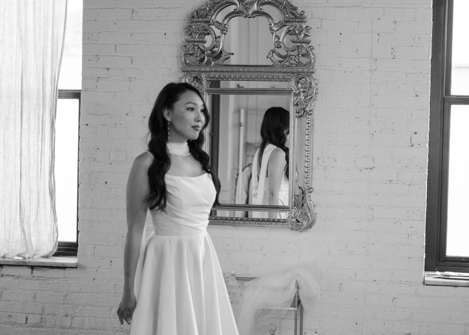 Woman in white wedding dress poses elegantly before ornate mirror in black and white photo.