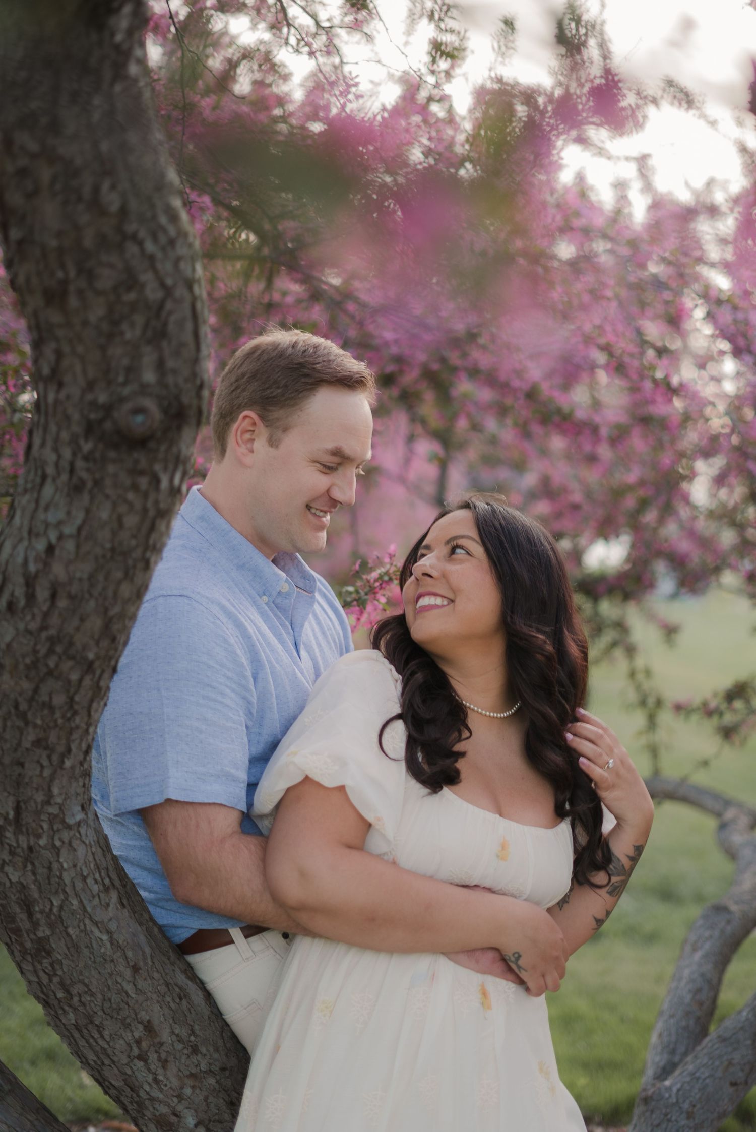 A couple embraces and smiles at each other near a tree with pink blossoms in a romantic outdoor engagement photo.