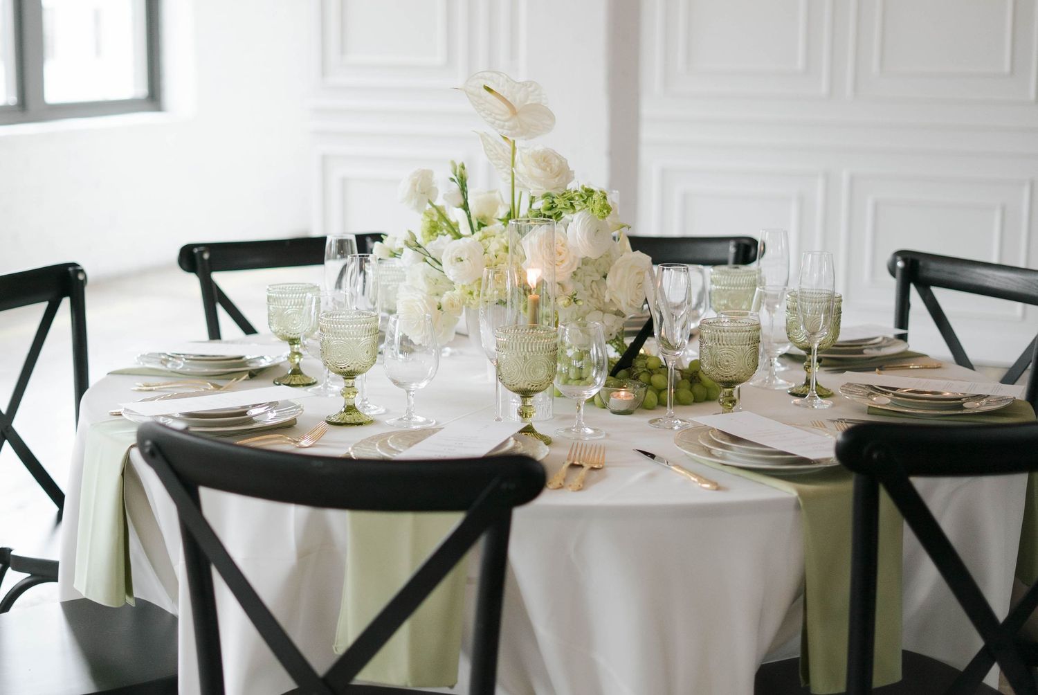 Elegant round wedding reception table with white floral centerpiece, black cross-back chairs, and sage green linens.