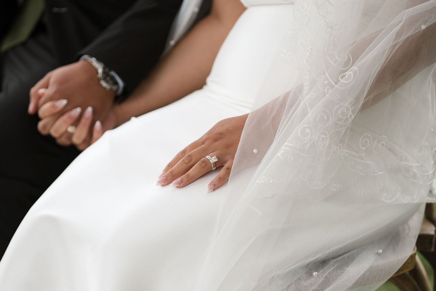 Close-up of bride's hand with diamond engagement ring resting on white wedding dress beside groom.