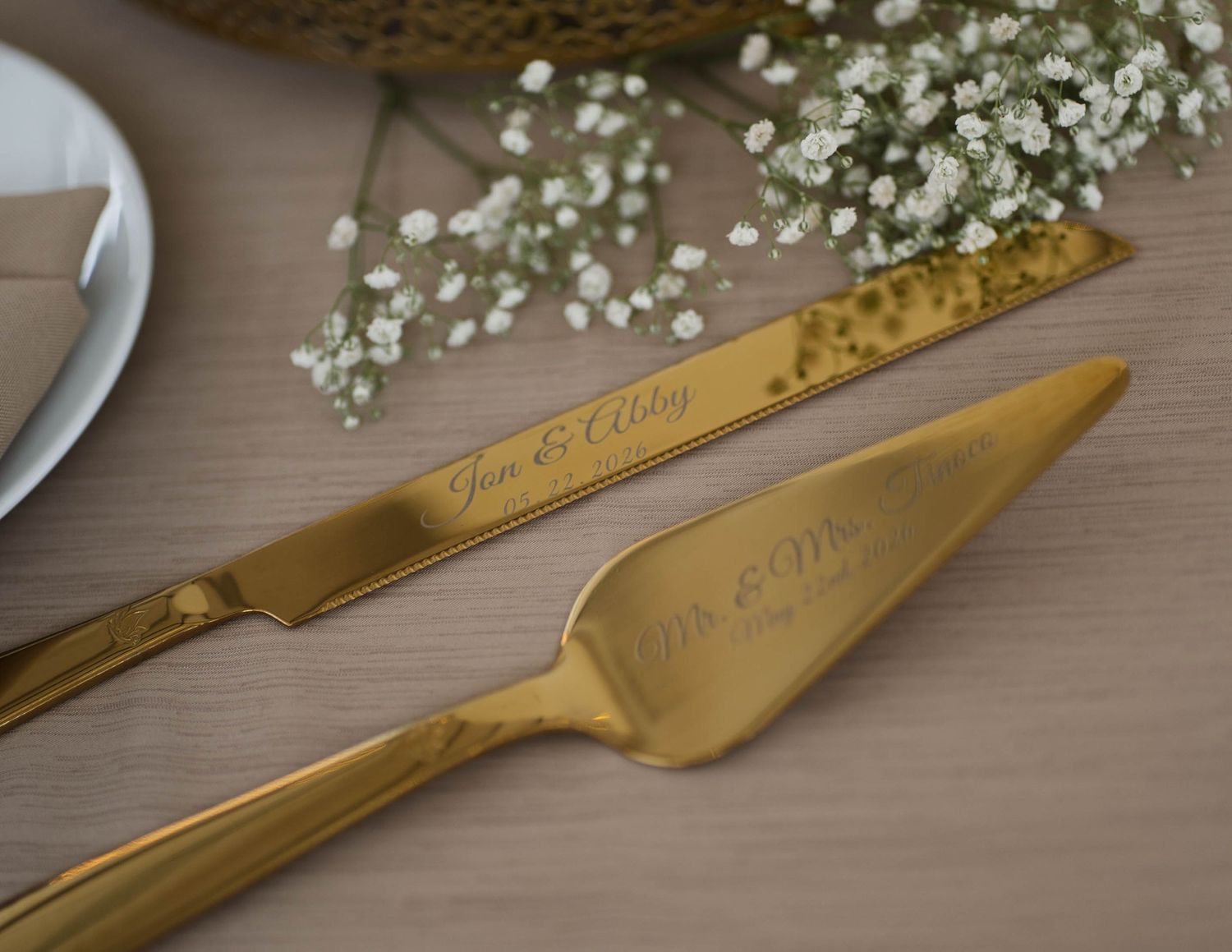 Engraved gold wedding cake cutting set with baby's breath flowers on wooden table.