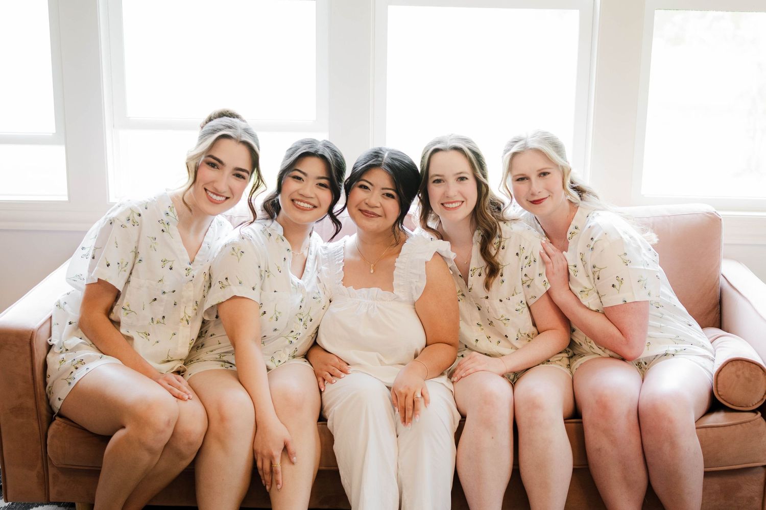 A bride poses with four bridesmaids in matching floral robes sitting on a pink sofa.