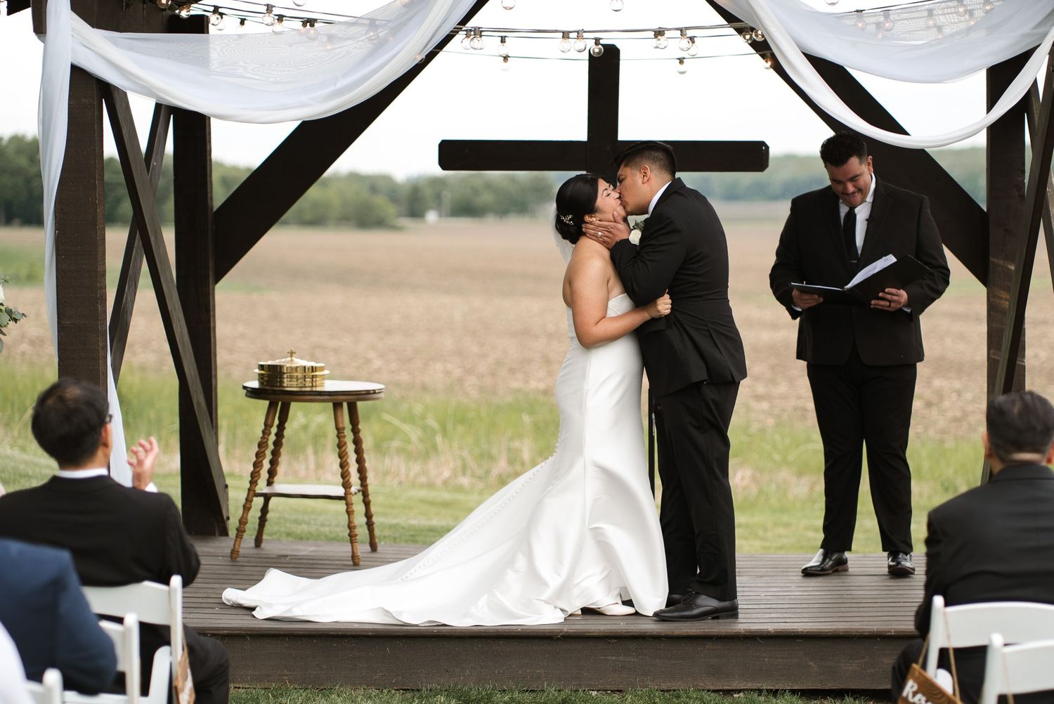 Bride and groom share first kiss at rustic outdoor wedding altar with cross backdrop and officiant.