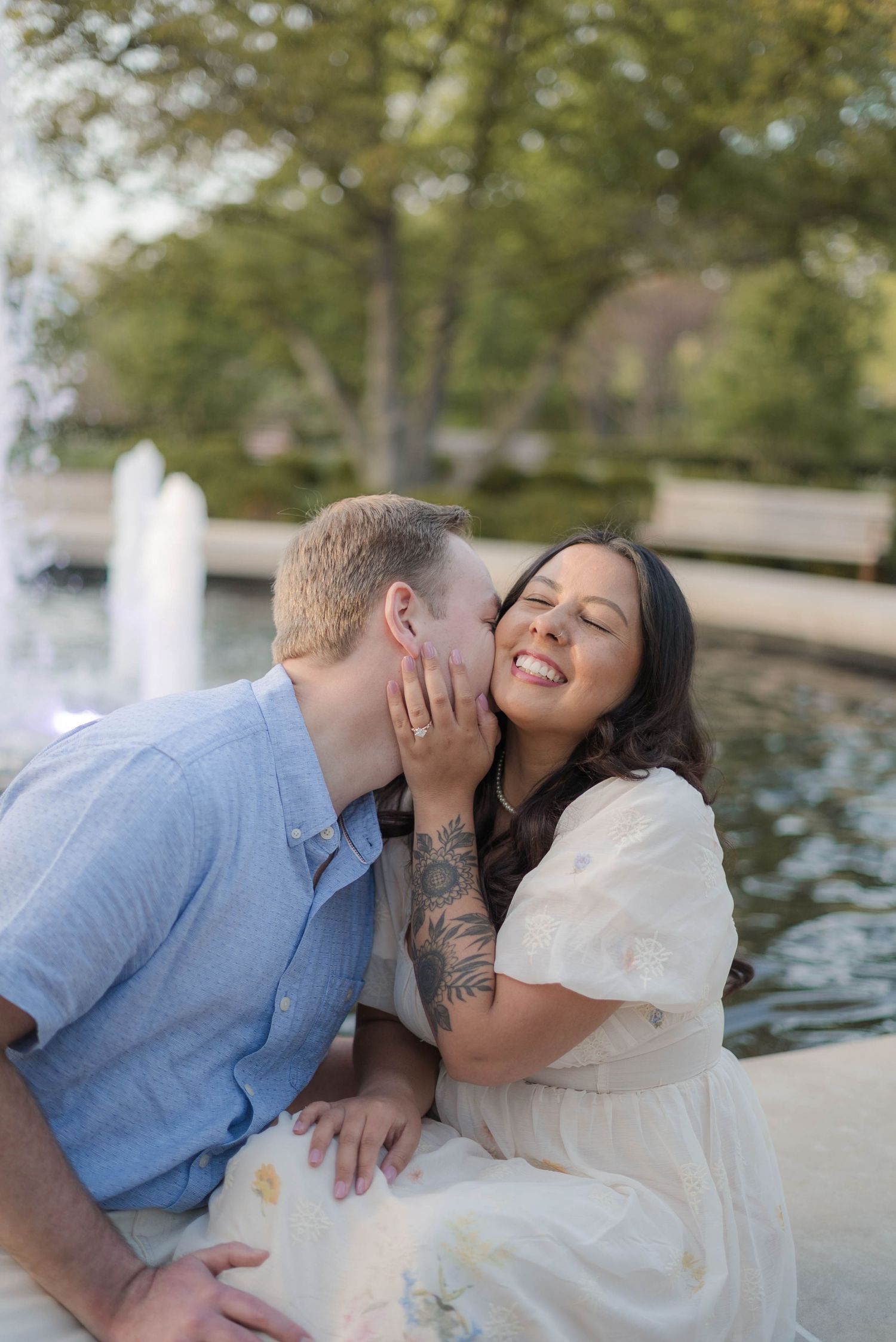 A couple shares a romantic kiss near a fountain, the woman smiling joyfully in a white dress with floral tattoos.