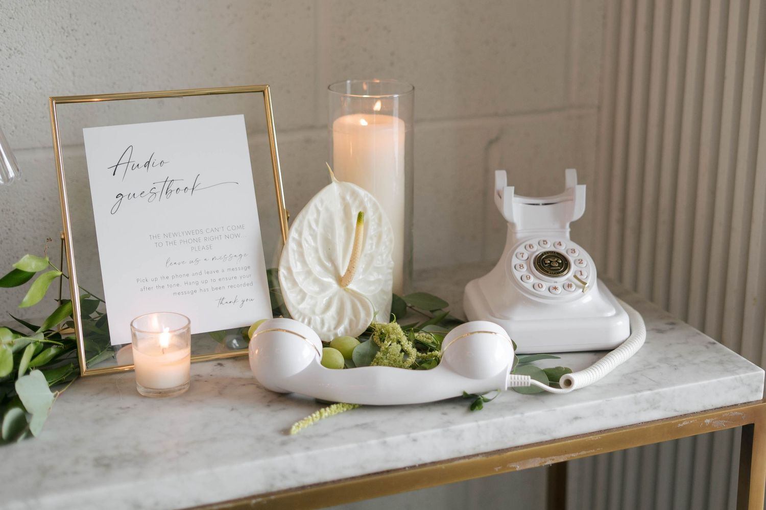 Elegant wedding audio guestbook display with white rotary phone, candles, greenery, and framed sign on marble table.