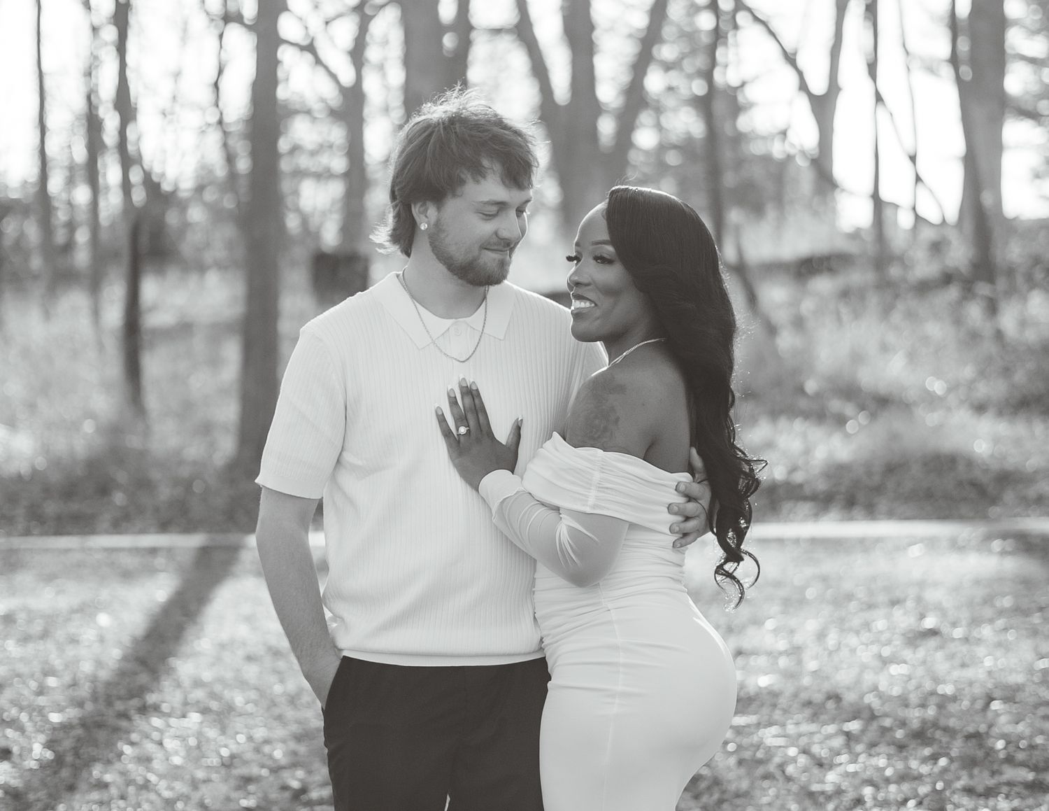 Black and white photo of a couple sharing a romantic moment outdoors among trees in a wooded setting.