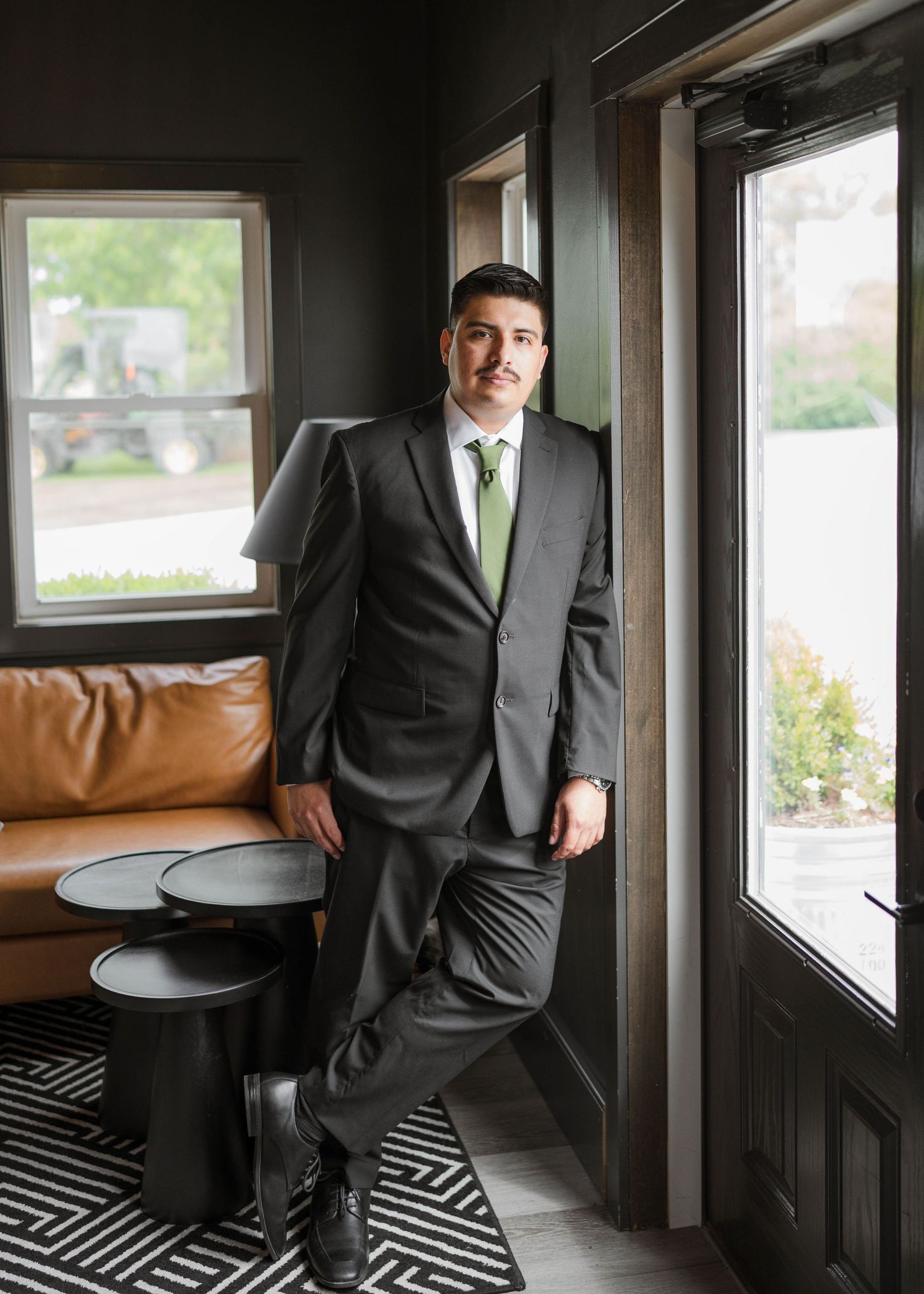 Groom in dark suit with green tie leaning against door frame in modern venue interior.