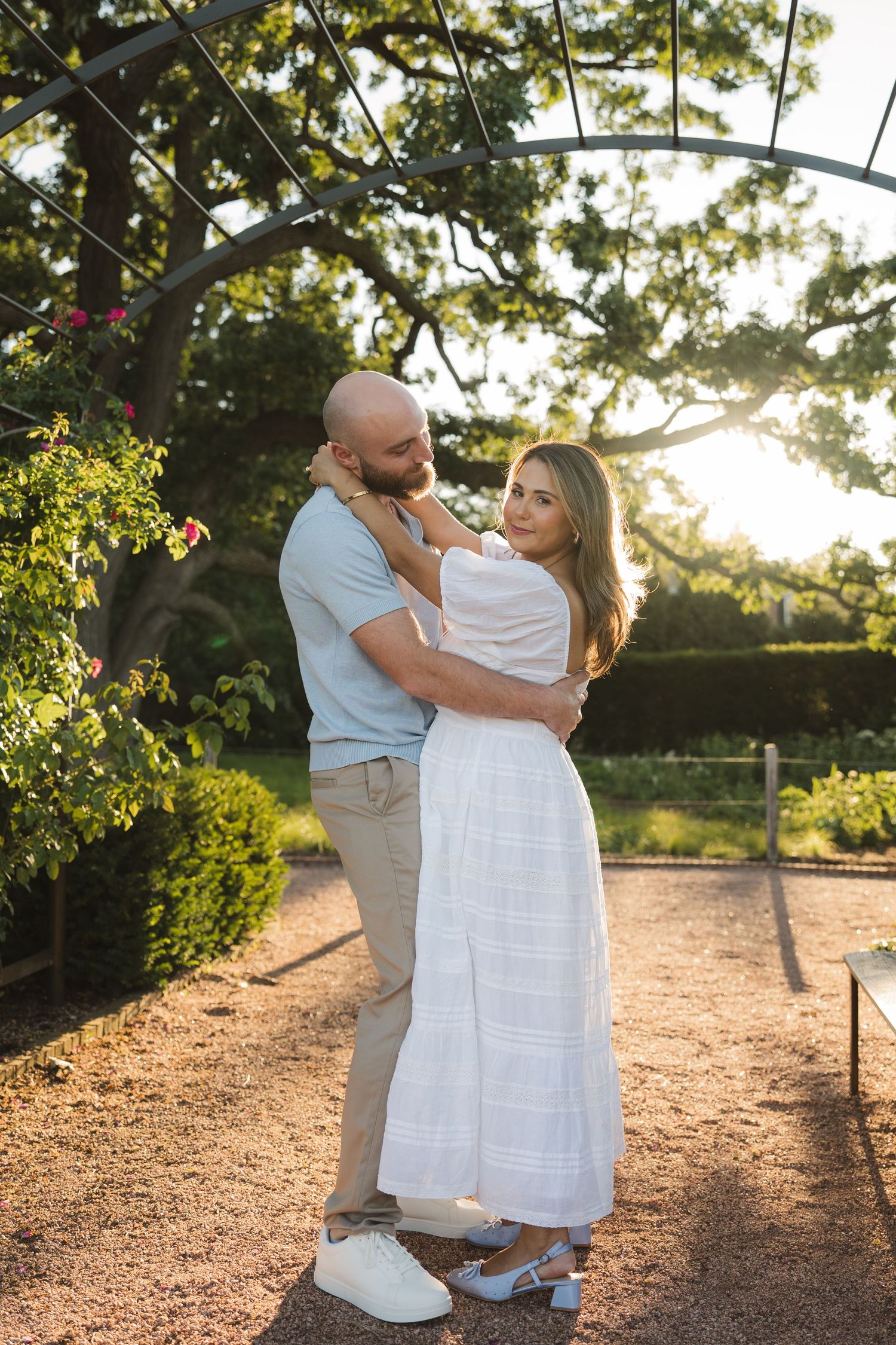 Couple embracing in a sunlit garden, woman in white dress and man in blue shirt, surrounded by lush greenery.