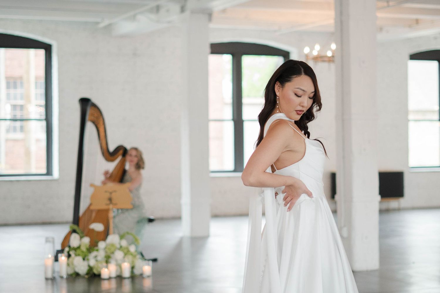 Bride in elegant white gown poses gracefully in bright studio with harpist performing in background.