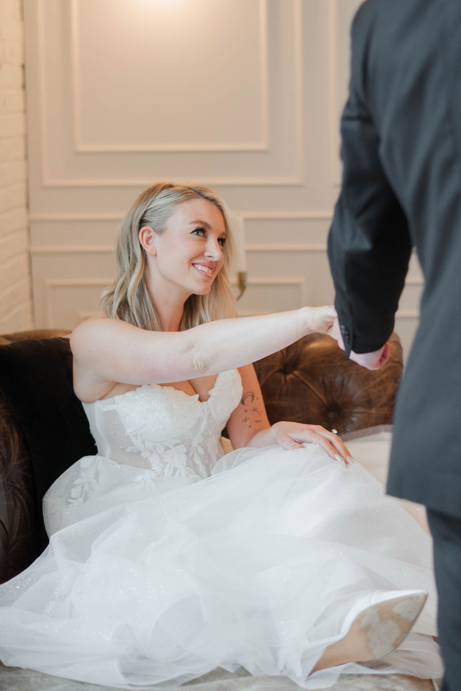 Smiling bride in white gown sits on leather sofa, reaching out to hold groom's hand on wedding day.