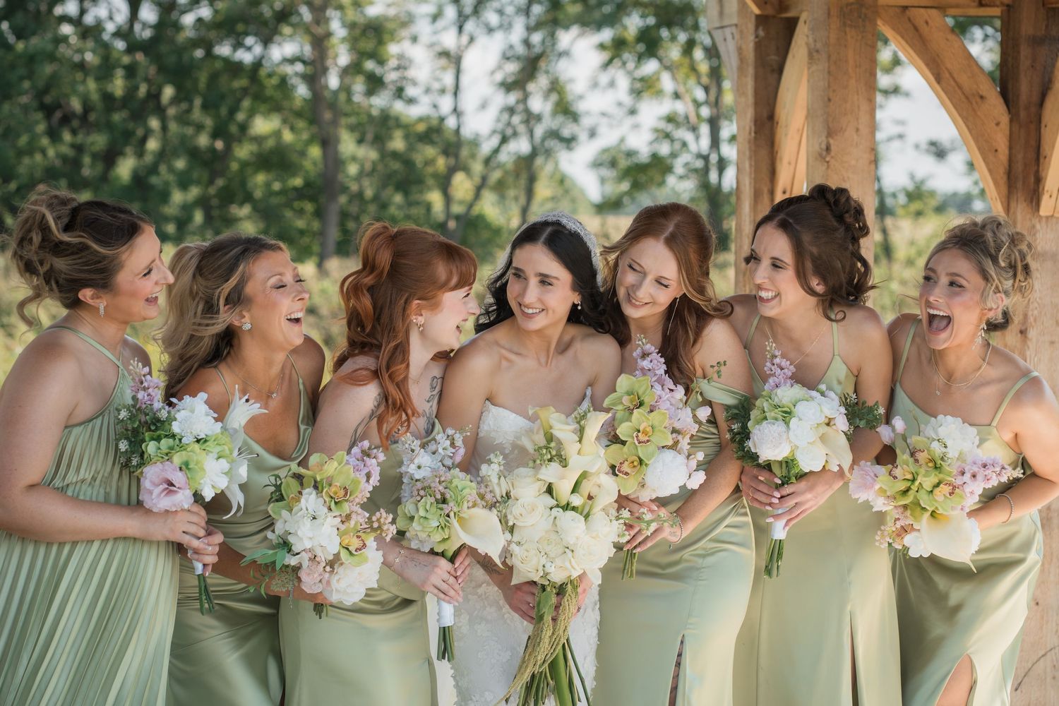 Bride and bridesmaids in sage green dresses laughing and holding floral bouquets outdoors at a rustic wedding venue.