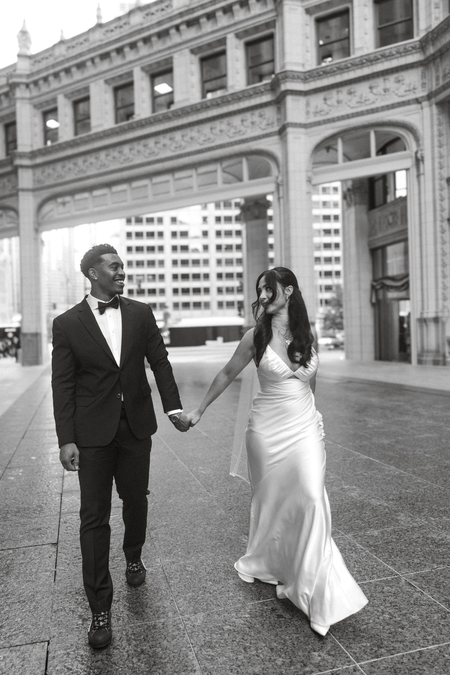 Black and white photo of a couple in wedding attire holding hands while walking in front of a grand urban building.