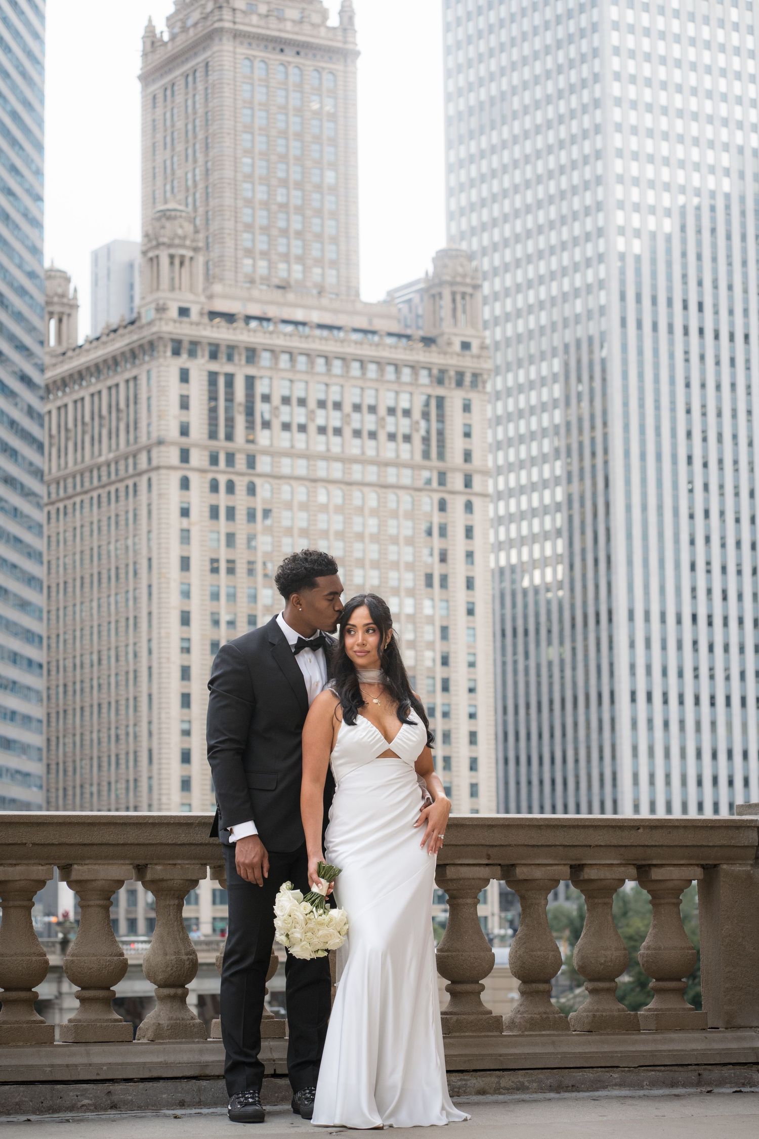 Elegant bride and groom posing on a balcony with Chicago skyscrapers in the background on their wedding day.