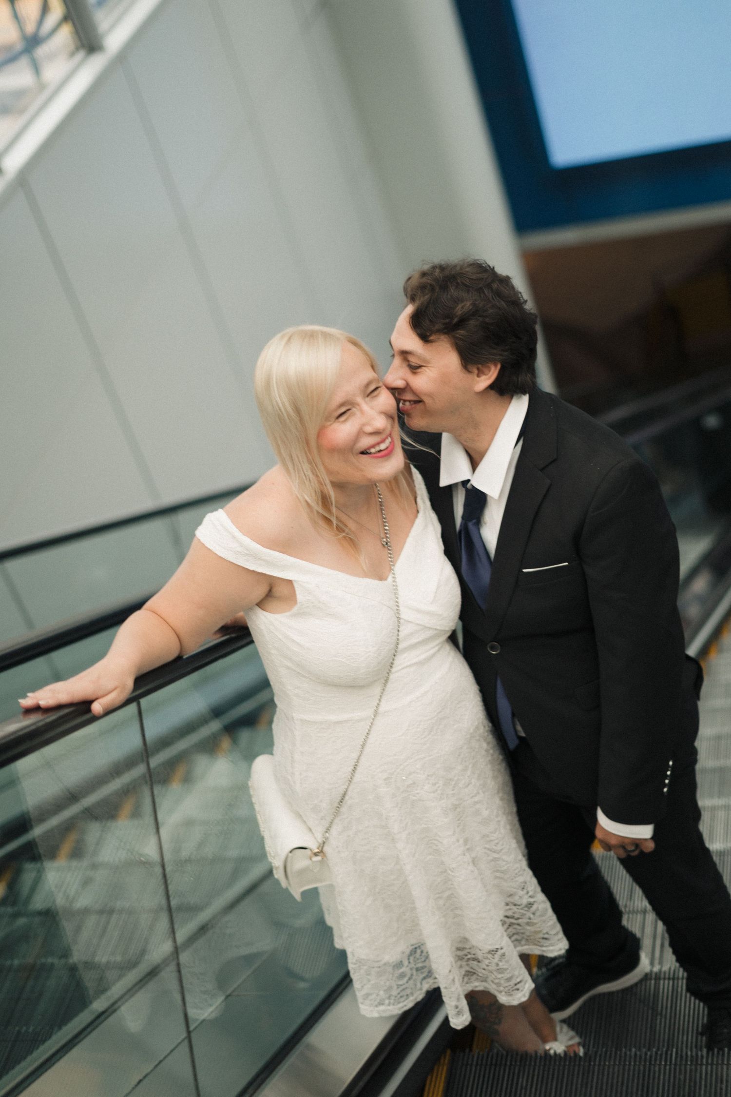 A smiling bride in a white lace dress and groom in a black suit share a tender moment on an escalator.
