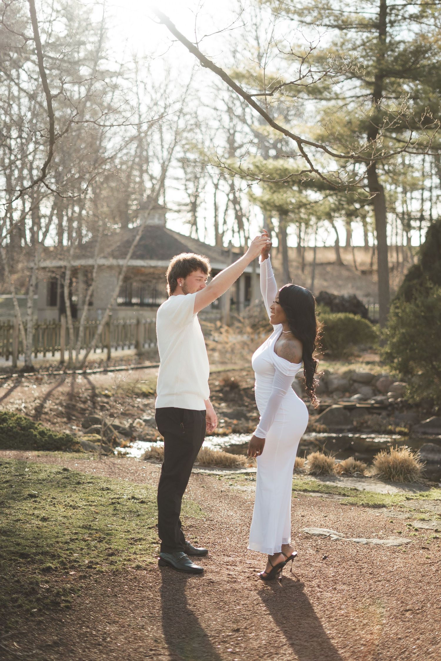 Couple dancing outdoors in a sunlit park, man in white tee and woman in white dress, surrounded by trees and a gazebo.