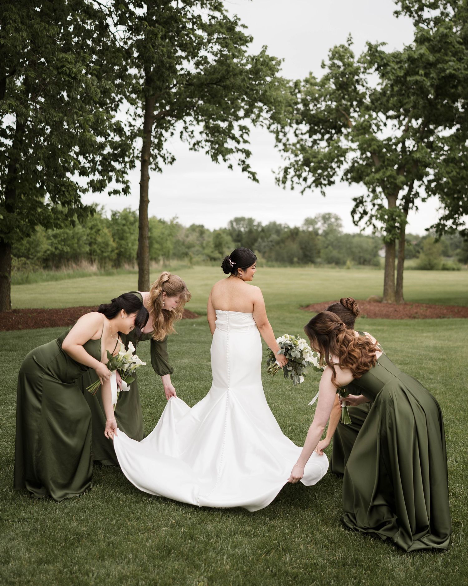 Bridesmaids in olive green gowns arranging the bride's white mermaid dress train on a lush green lawn.