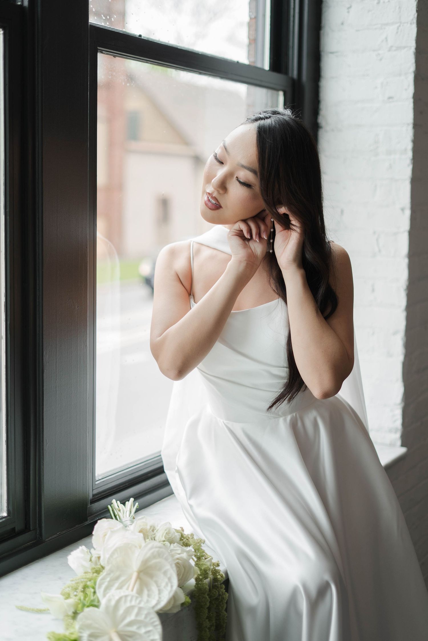 Elegant bride in white satin gown adjusting earring by window with white floral bouquet in foreground.