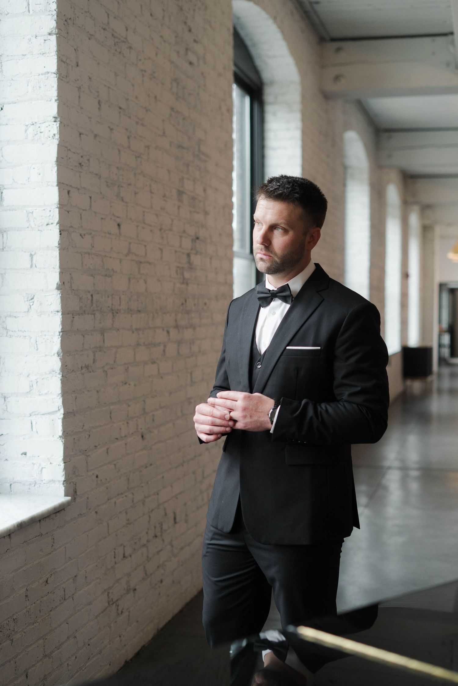 A dapper groom in a black tuxedo with bow tie stands confidently in an elegant venue with exposed brick walls.