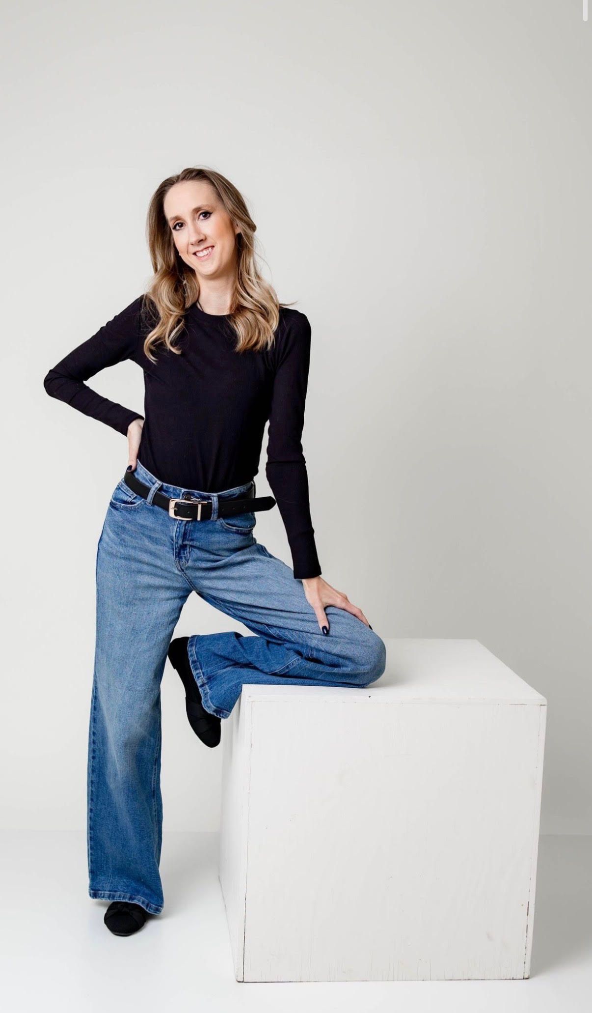 Woman in black long-sleeve top and wide-leg jeans posing confidently against a white background.