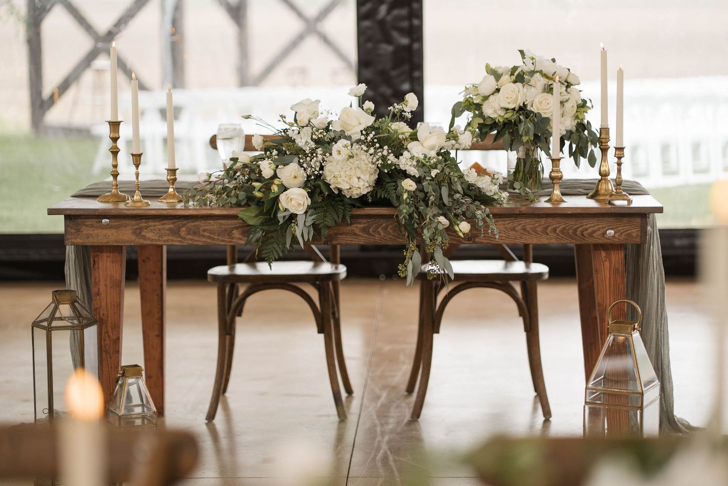 Rustic wooden sweetheart table adorned with white floral arrangements, greenery, and gold candlesticks for a wedding reception.