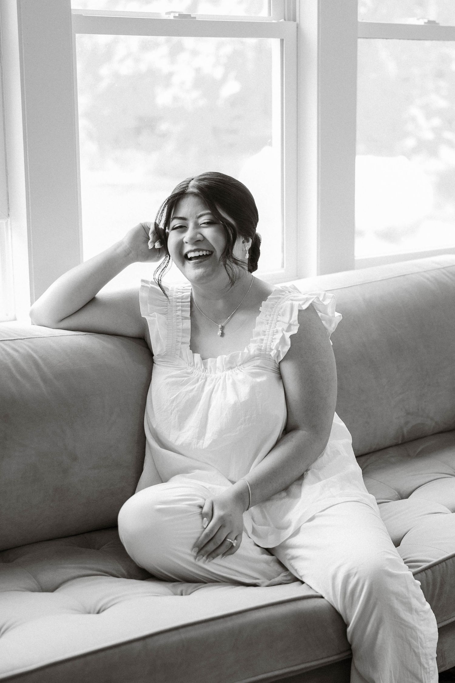 Pregnant woman in white dress laughing joyfully on a couch by a bright window in black and white.