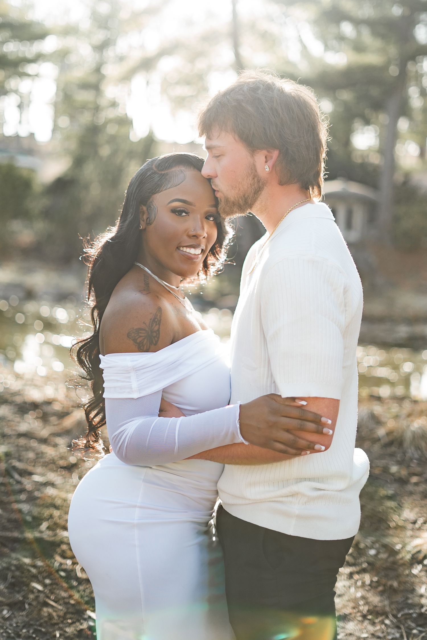 A couple embraces outdoors, woman in white off-shoulder dress smiling at camera, man kissing her temple in sunlit setting.