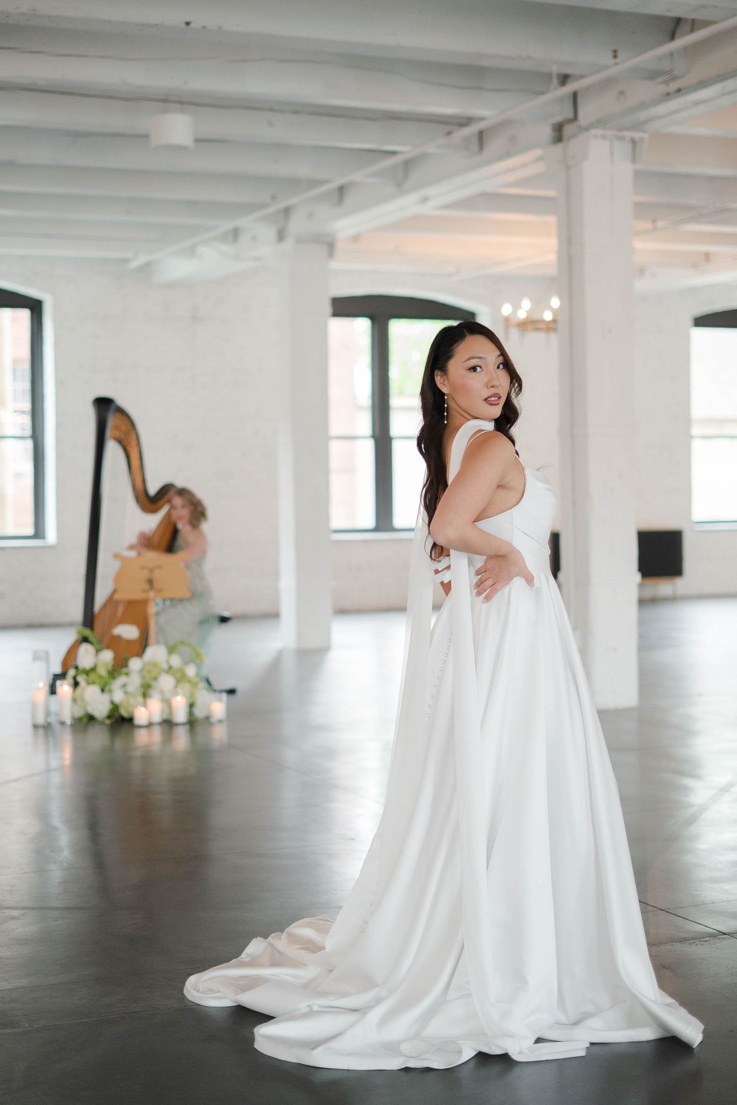 Bride in elegant white satin gown poses in bright venue with harpist playing in background.
