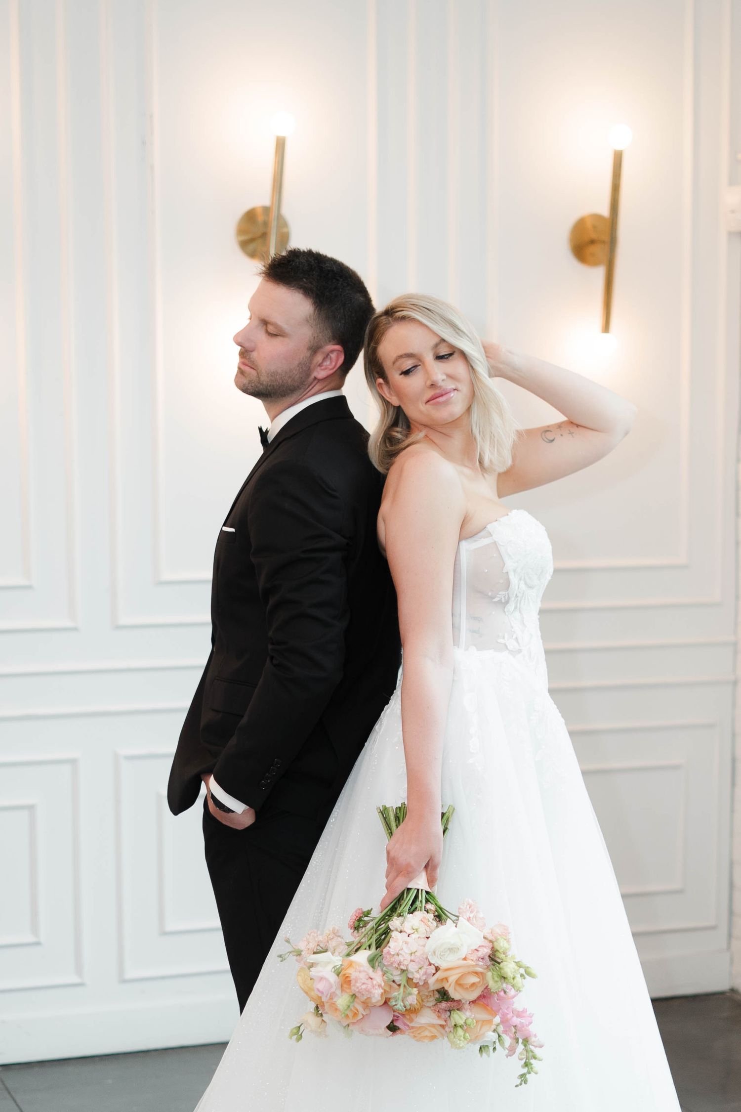 Bride and groom posing back-to-back in elegant venue, bride holding pink and white bouquet in strapless gown.