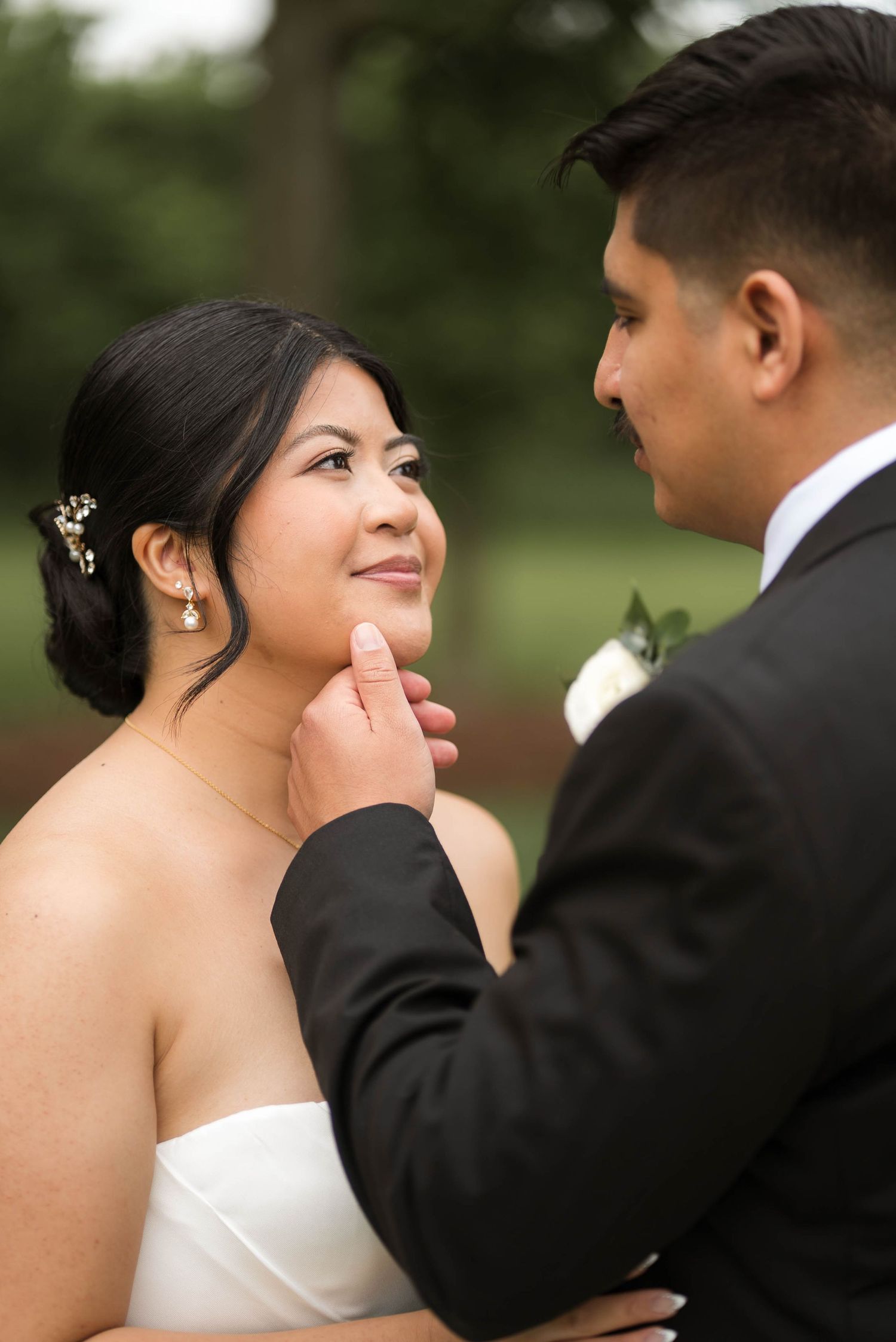 Groom tenderly lifts bride's chin as she gazes up smiling in romantic outdoor wedding portrait.