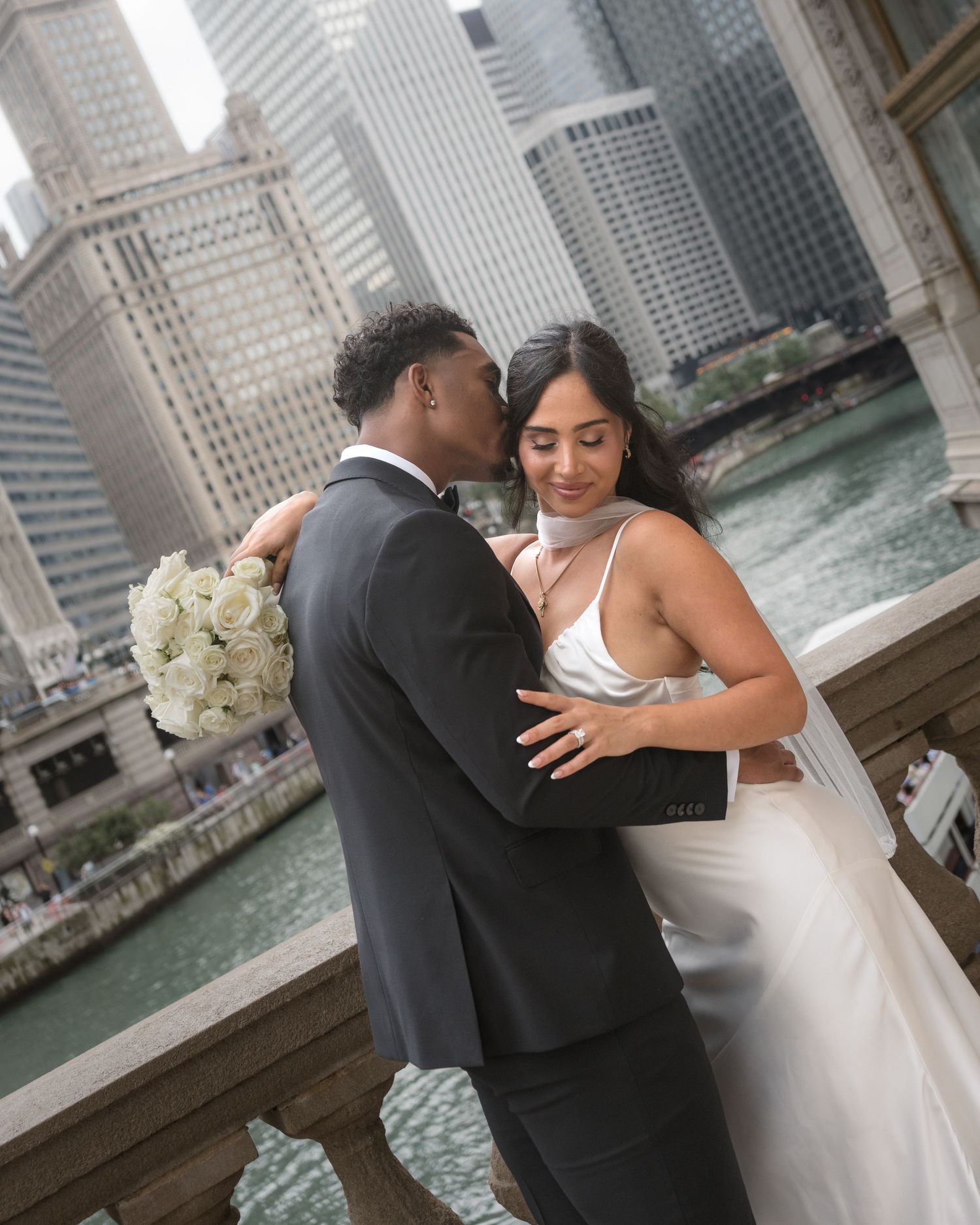 Couple in wedding attire embracing on Chicago Riverwalk bridge, groom kissing bride holding white roses bouquet.