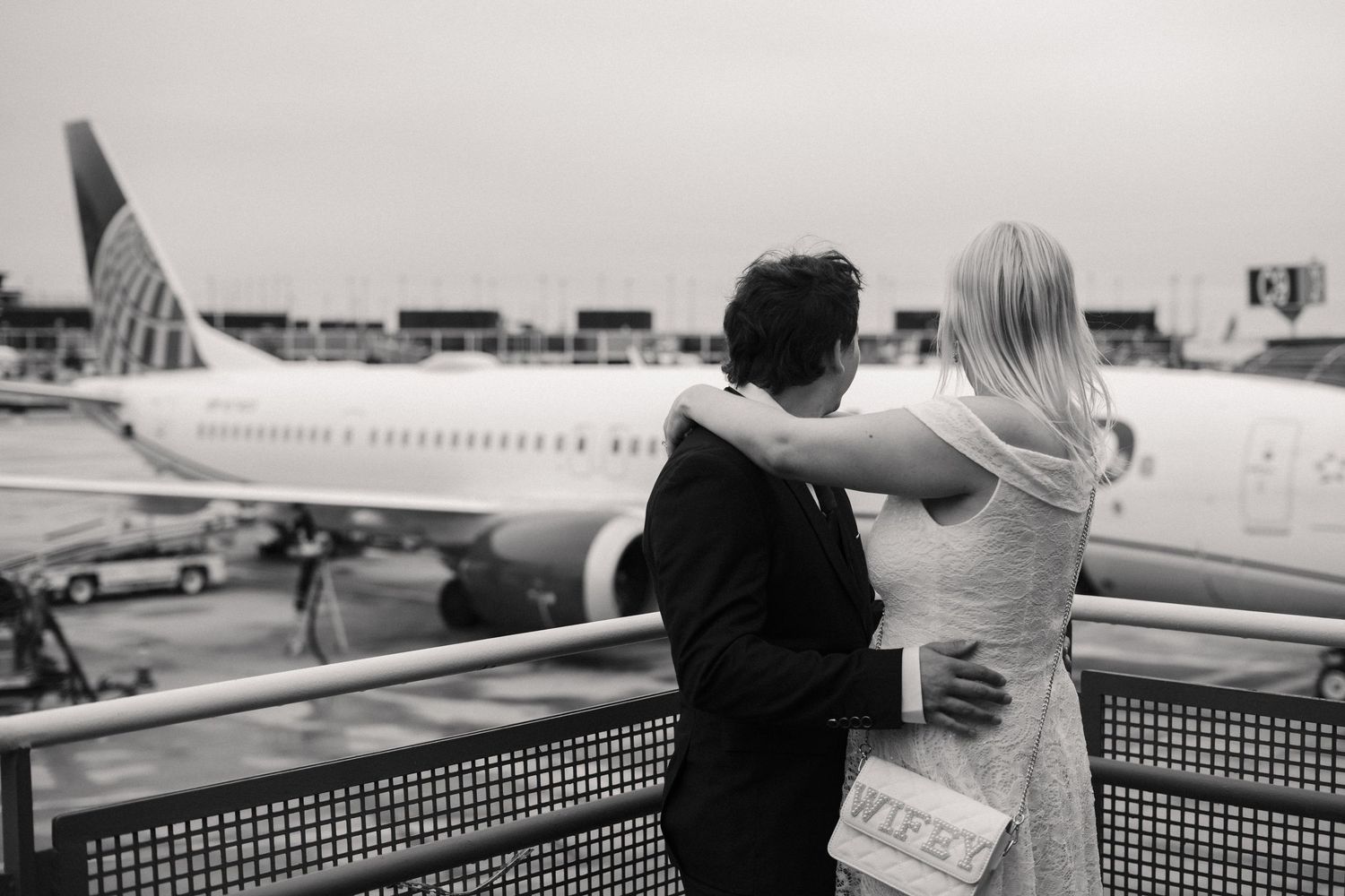 Couple embracing at airport terminal window watching airplane on tarmac in black and white photo.