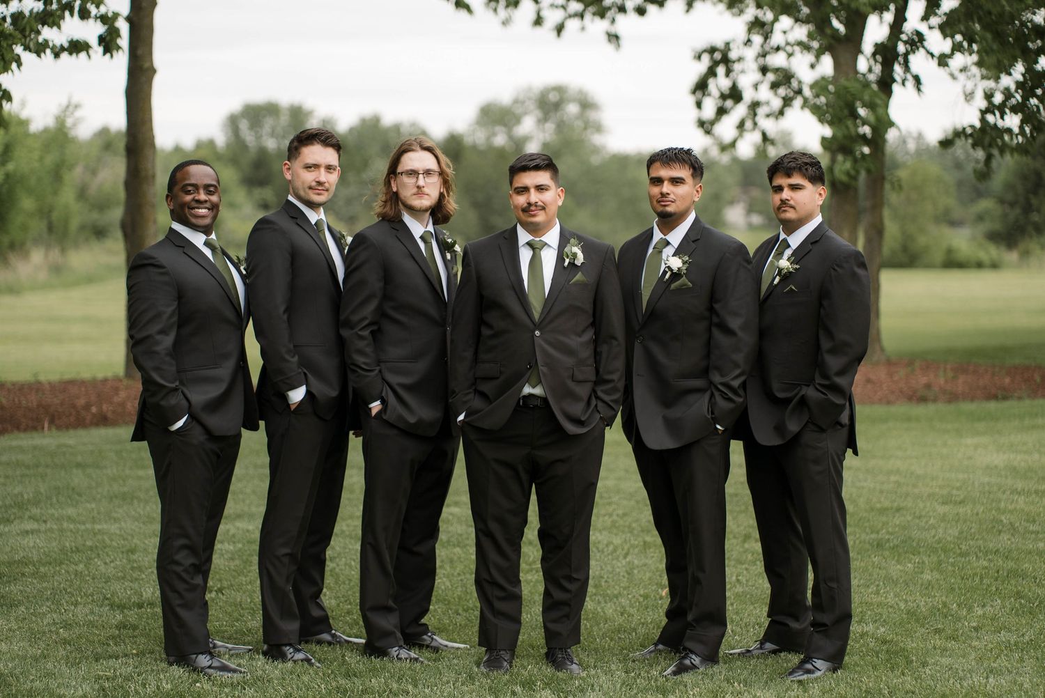 Six groomsmen in black suits with green ties posing outdoors on a grassy lawn.