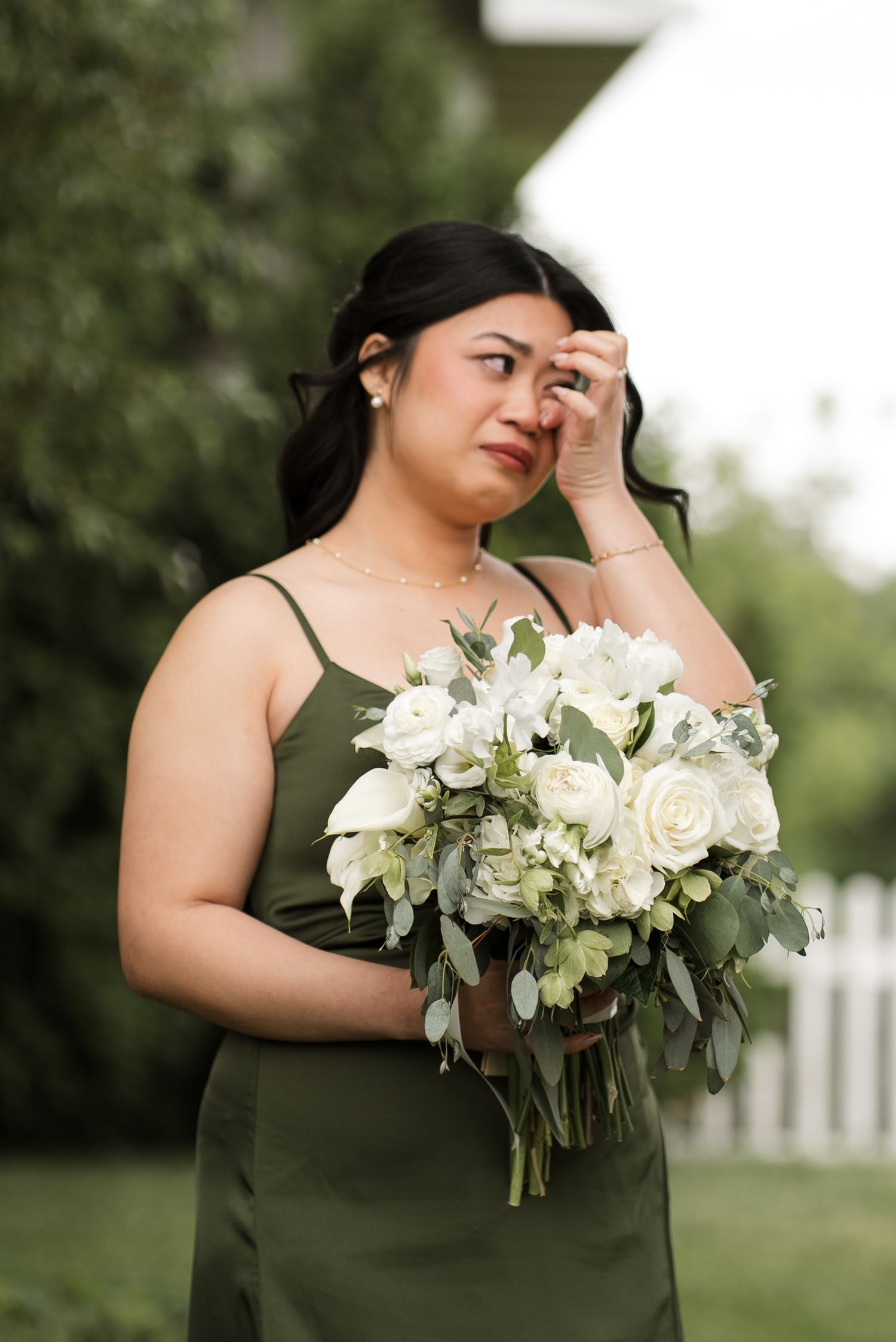 Bridesmaid in olive green dress holding white rose bouquet, wiping tear at outdoor wedding ceremony.