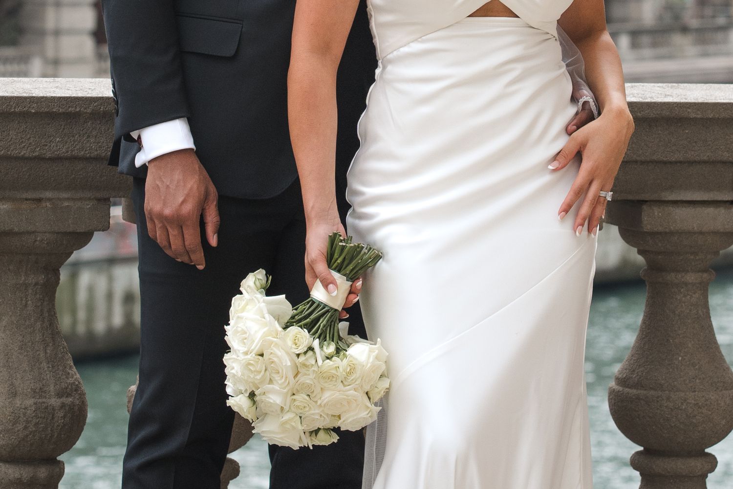 A bride in a white gown and groom in a black suit hold a white rose bouquet at their wedding.