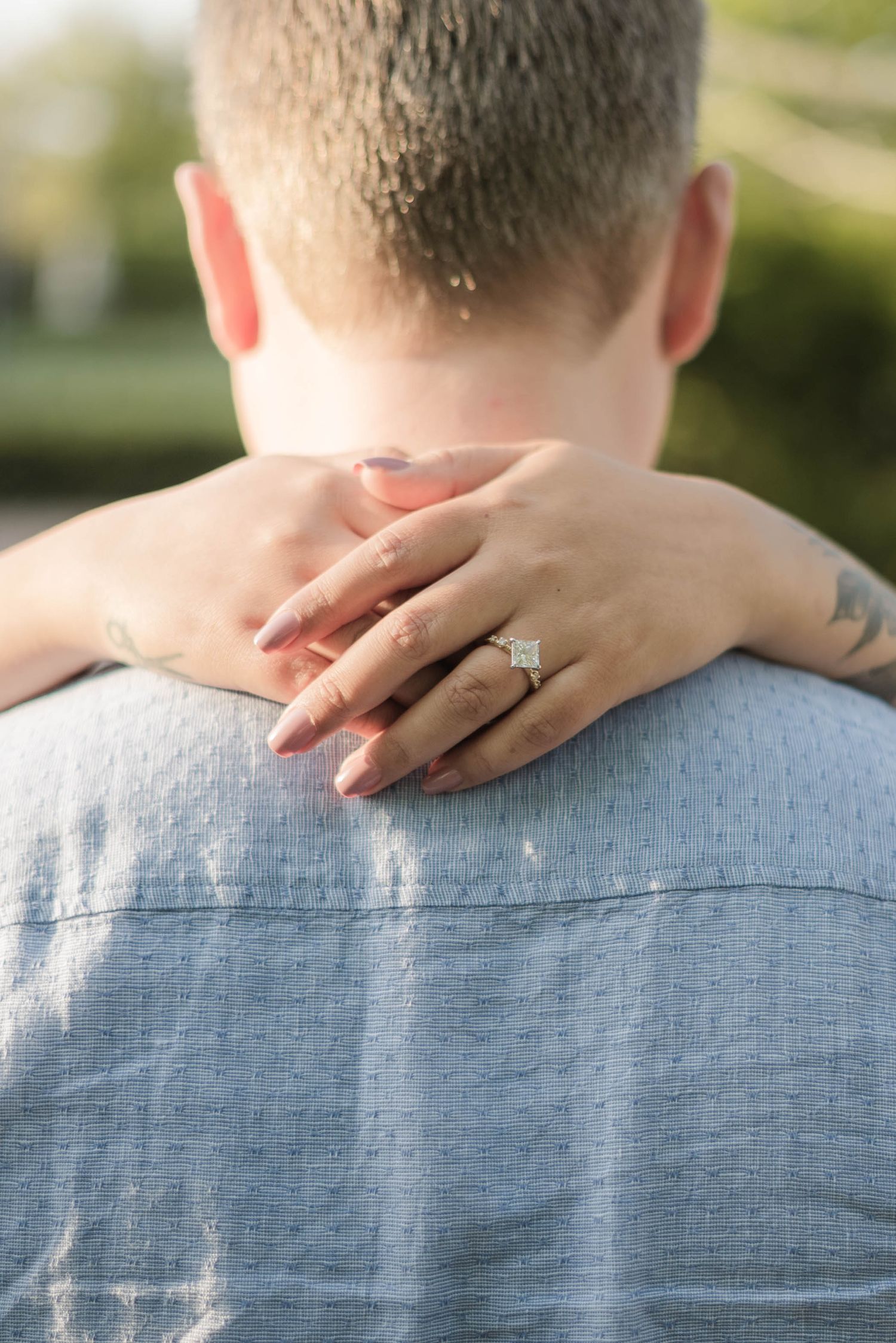 Woman with engagement ring rests hands on mans shoulders from behind, captured in soft natural light.
