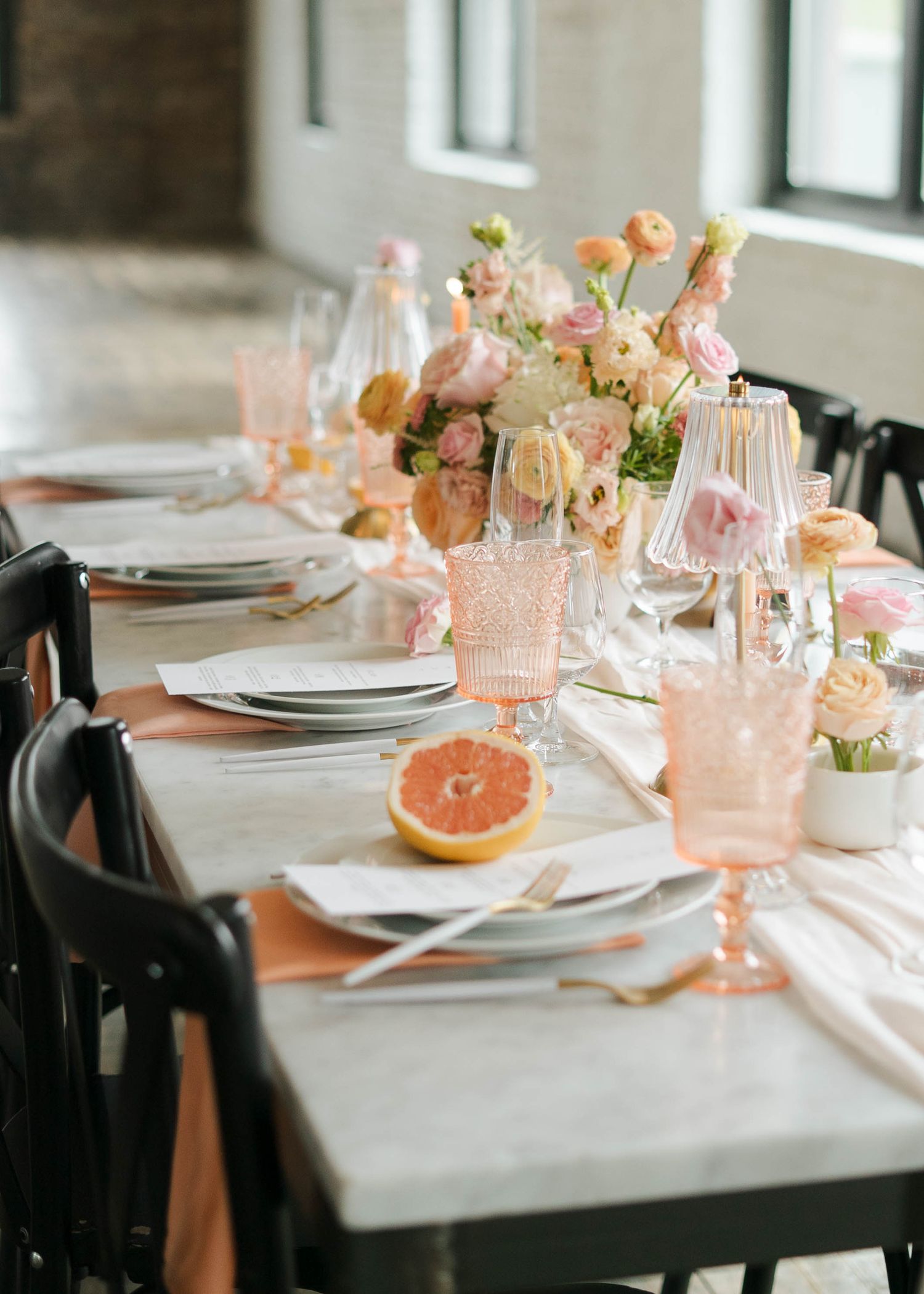 Elegant wedding tablescape with peach florals, pink glassware, marble table, black chairs, and a halved grapefruit centerpiece accent.