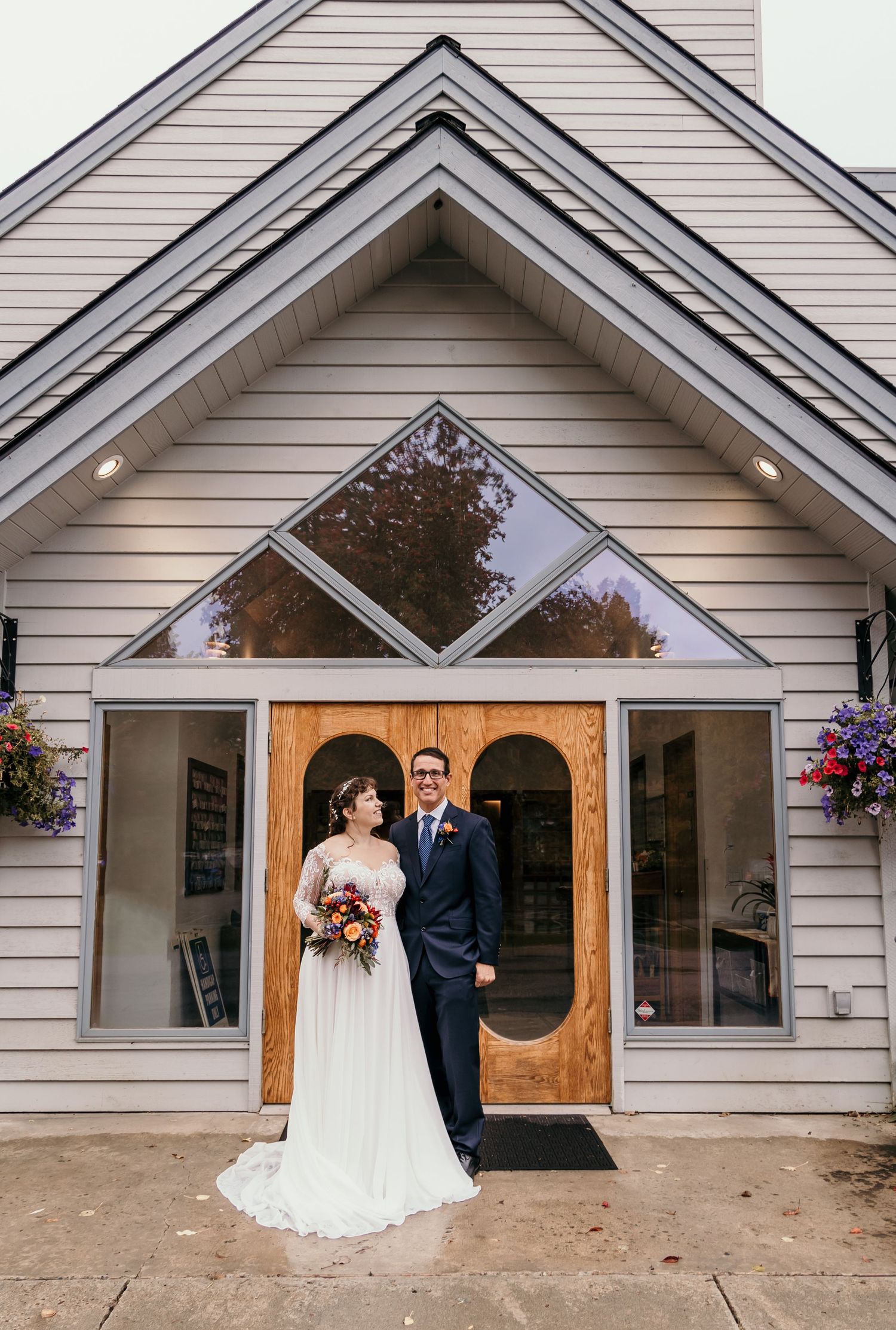 Bride and groom pose in front of a chapel with wooden arched doors and triangular glass window detail.