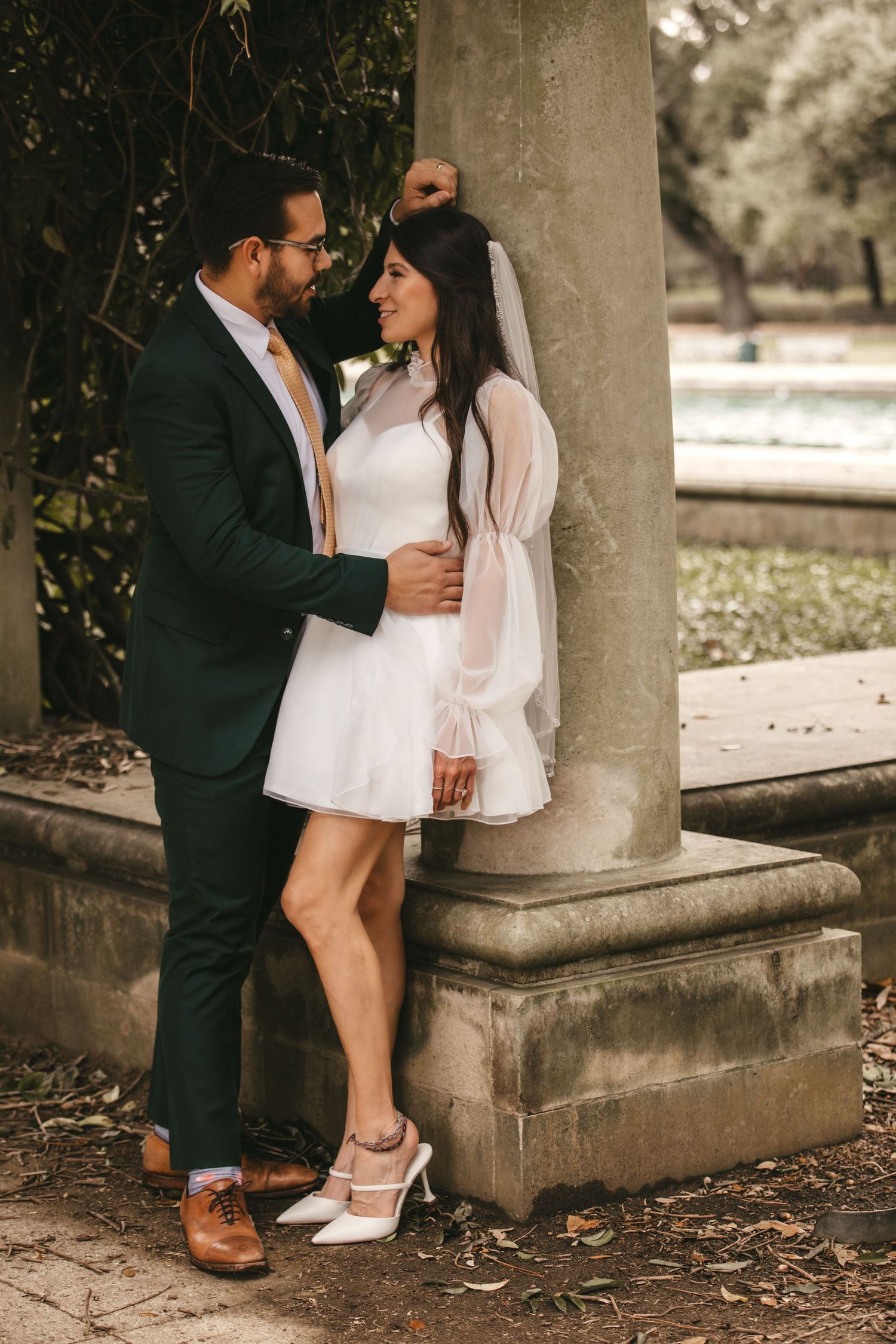Wedding couple embraces by stone column outdoors, bride in mini white dress with veil and groom in dark green suit.