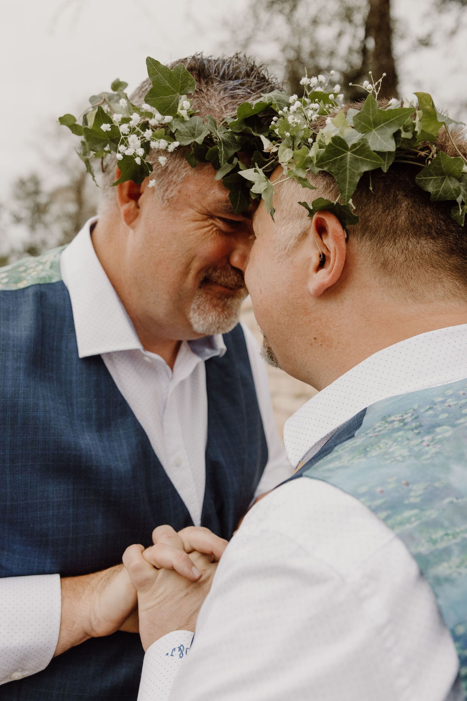 Two grooms in floral crowns share an intimate moment holding hands at their wedding ceremony.