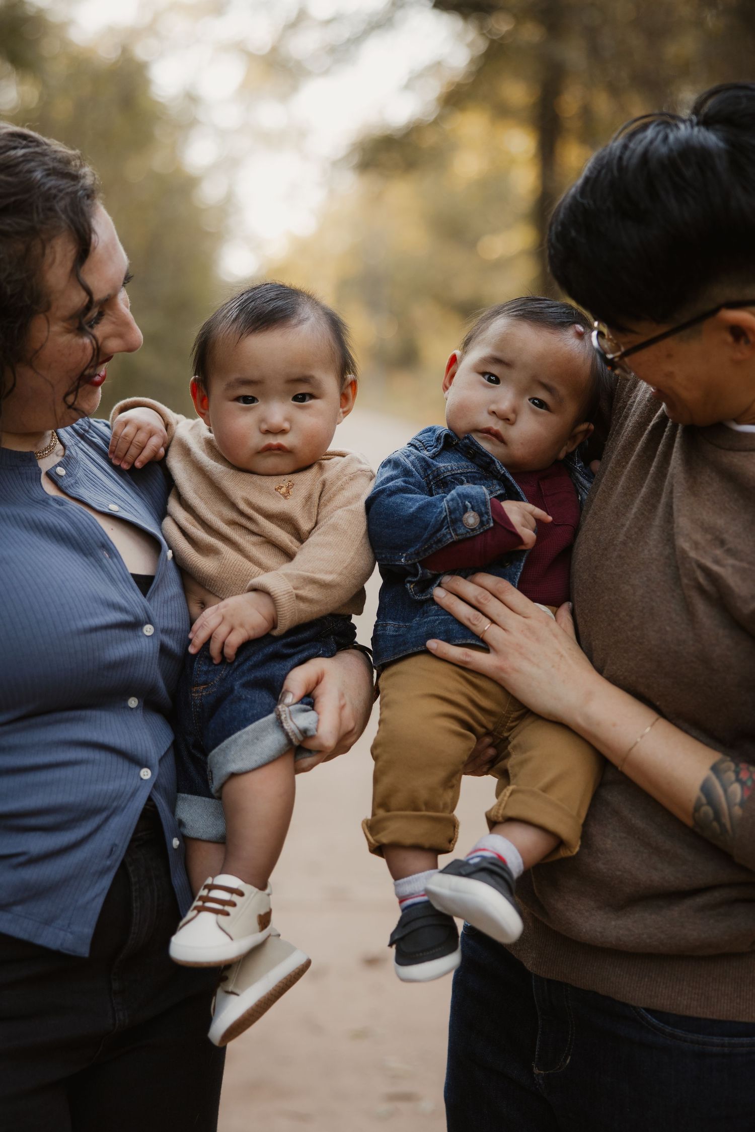 Two parents holding twin baby boys face-to-face on a sunlit autumn path in the park.