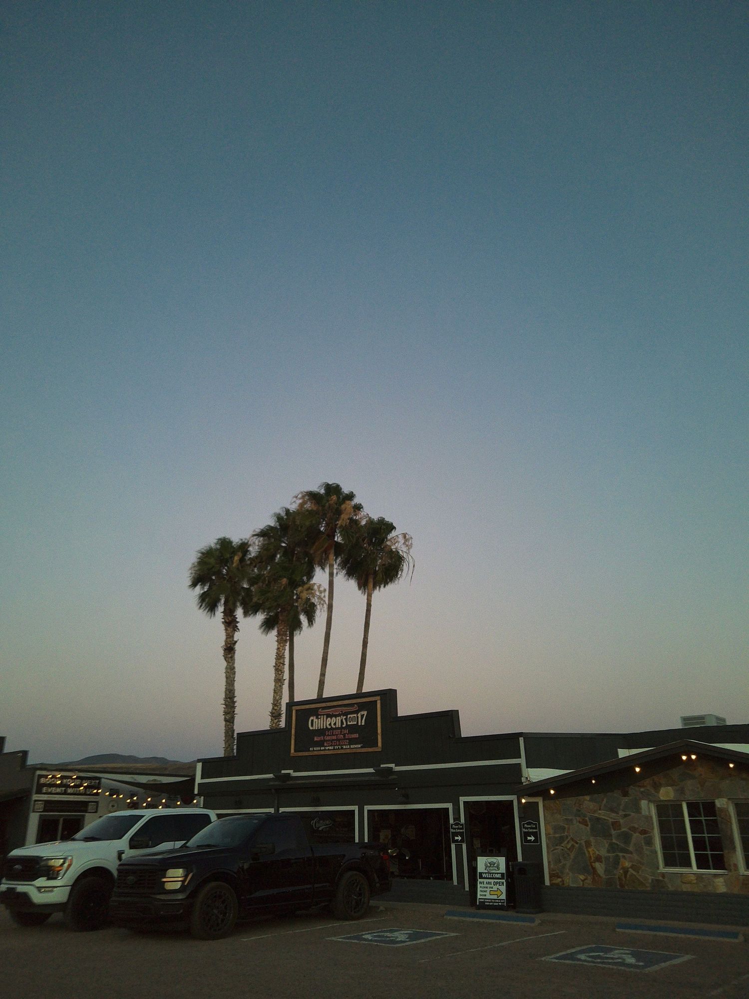 Palm trees silhouette against a twilight sky above a dark restaurant building with string lights in a parking lot.