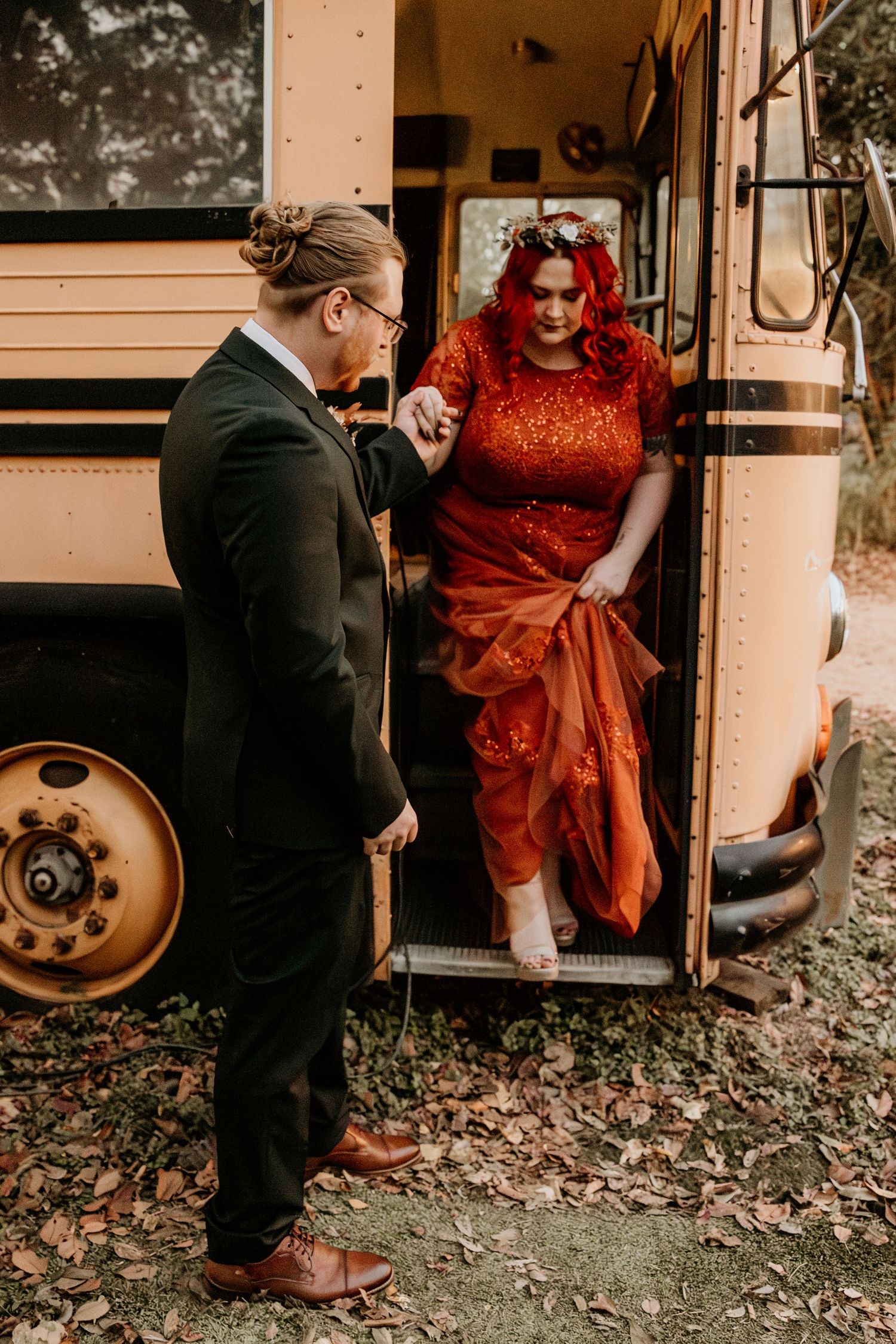 Groom helps bride in red sequin gown step off vintage yellow school bus on wedding day.